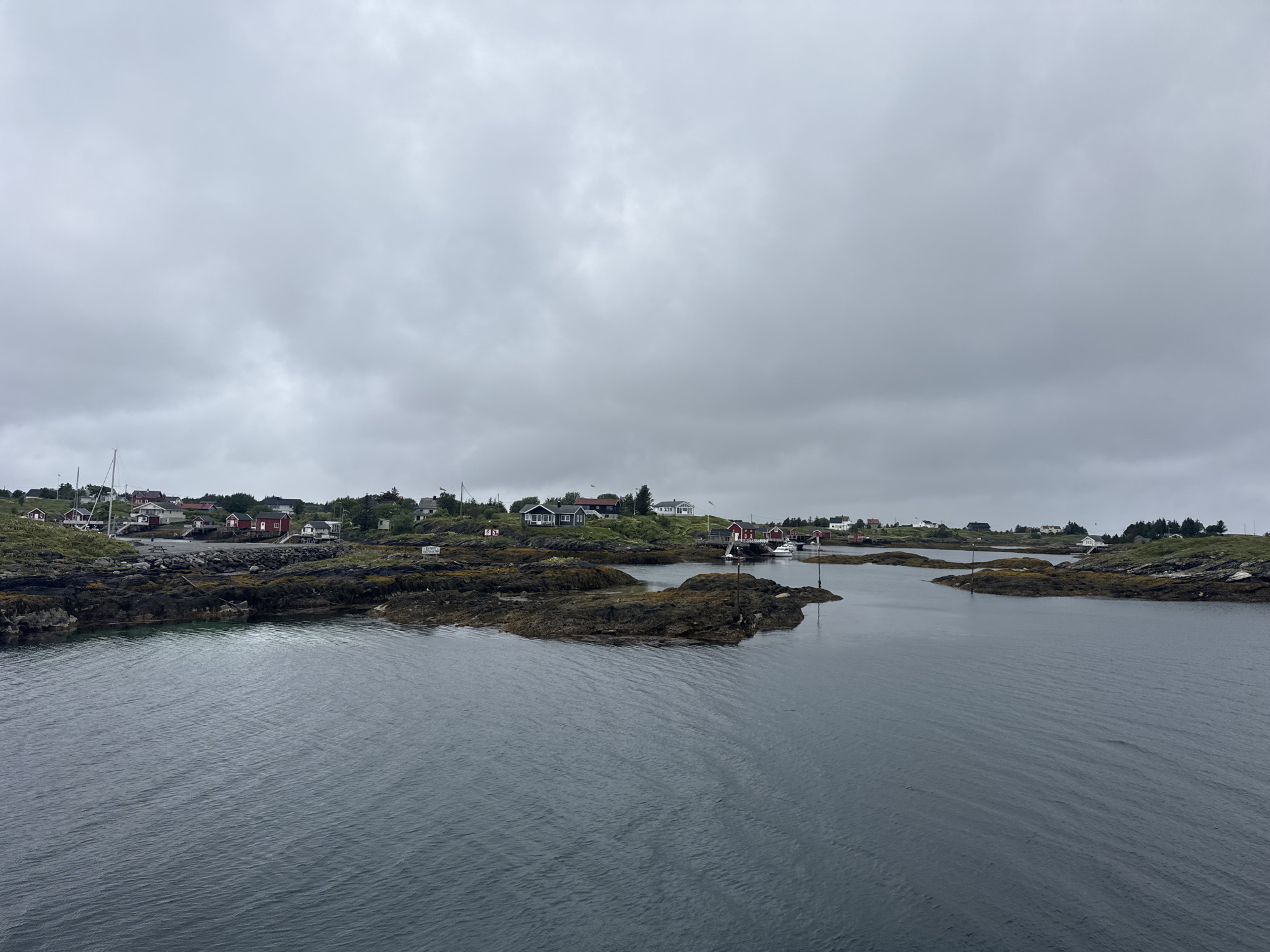The image depicts a panoramic view of a coastal settlement on Sleneset, Norway, under an overcast sky. In the foreground, dark, rippling water stretches across the frame, leading towards a cluster of rocky islands dotted with sparse vegetation. These islands are intricately connected to the mainland, forming a natural harbor or bay. On the mainland and the islands, a small village of red and white houses is visible, nestled amongst the rocky terrain and green hills. Several boats are anchored in the calm waters, and a few small sailing vessels are present. The architecture of the houses suggests a traditional Scandinavian coastal style. The overall atmosphere is serene and maritime, indicative of a quiet fishing village or a small community by the sea. There are no people visible in the image.