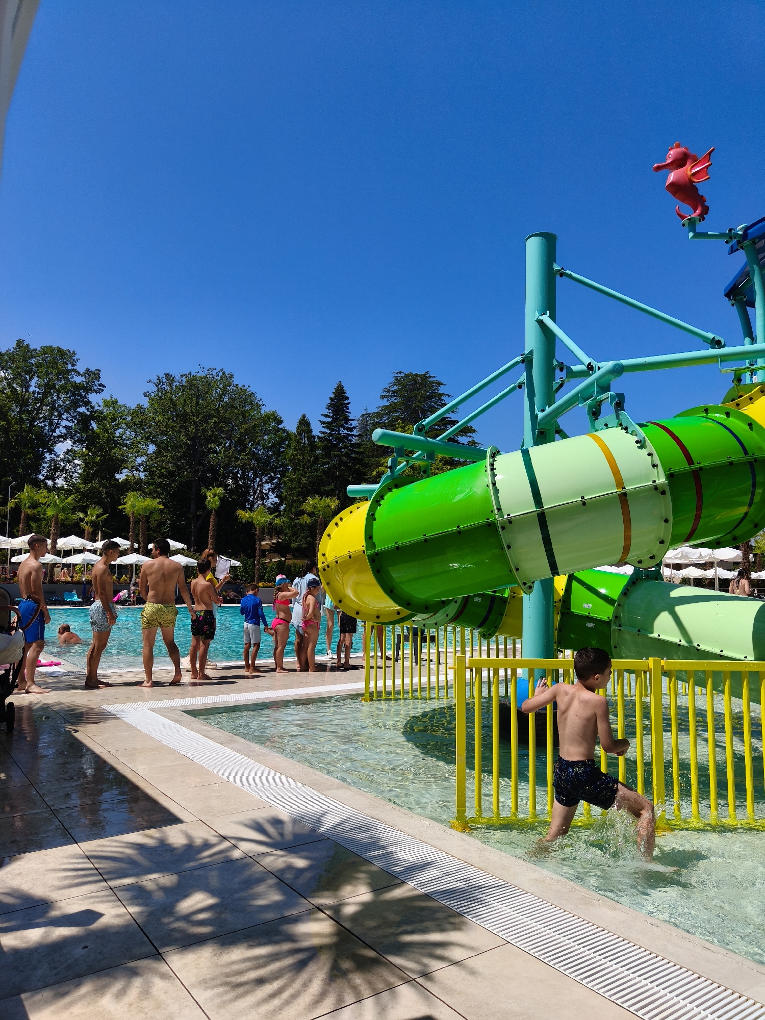 The image depicts a sunny day at a water park in Kiten, Bulgaria. A bright blue sky dominates the upper portion of the frame, with a red seahorse-shaped decoration visible on a turquoise structure of a waterslide. Below, a large waterslide complex with green and yellow tubes is a prominent feature. In the foreground, a young boy wearing dark patterned swim trunks is running through the shallow end of a pool, splashing water. Several other people, including children and adults, are gathered around the pool, some in swimsuits, and appear to be waiting in line or relaxing. Palm trees and lush green trees line the background, with white umbrellas suggesting a seating area. The pool water is clear and inviting, and the poolside area is paved with light-colored tiles, some of which reflect the sunlight and palm tree shadows. The overall scene conveys a sense of leisure and summer fun.