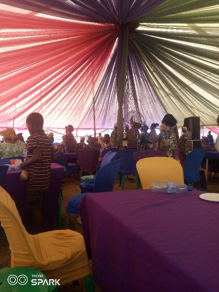 The image shows an event taking place under a large tent with colorful fabric draped from the center, creating a starburst effect. The fabric is predominantly red, purple, and green. The event appears to be in Oka-Akoko, Nigeria, based on contextual knowledge. Several people are seated at tables covered in purple tablecloths. The chairs have covers of various colors including yellow and blue. A child in a striped shirt is standing near one of the tables. In the background, there are more people and what appears to be a speaker. There is a plastic bottle and a plate on the table in the foreground. A fan is visible in the background, among the other people. The lighting is somewhat dim. The bottom of the image has the watermark of a 'TECNNO SPARK' phone.