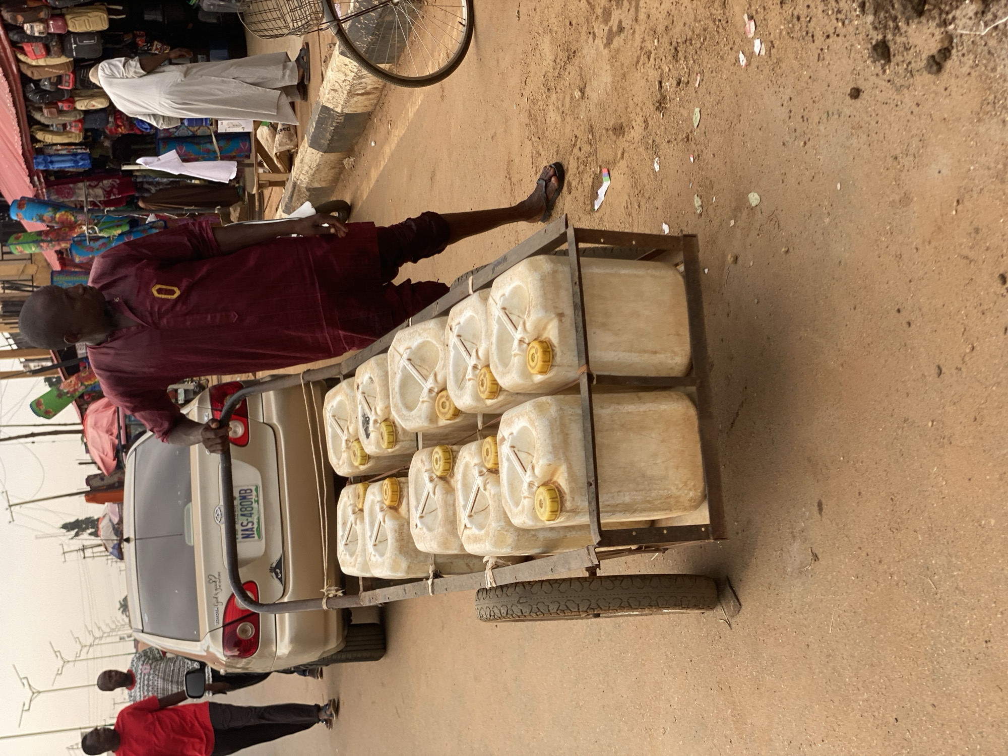A street scene in what is likely Kafanchan B, Nigeria, displays a man pulling a hand-pulled cart loaded with twelve white plastic jerrycans along the road. The road is dusty and appears unpaved in sections. The man wears a maroon outfit and sandals. A beige sedan, with license plate NAS-480MB, is beside the cart. The rear of the car is visible. The background includes street vendors displaying goods under tents. Other people are walking nearby. Overhead power lines are visible.