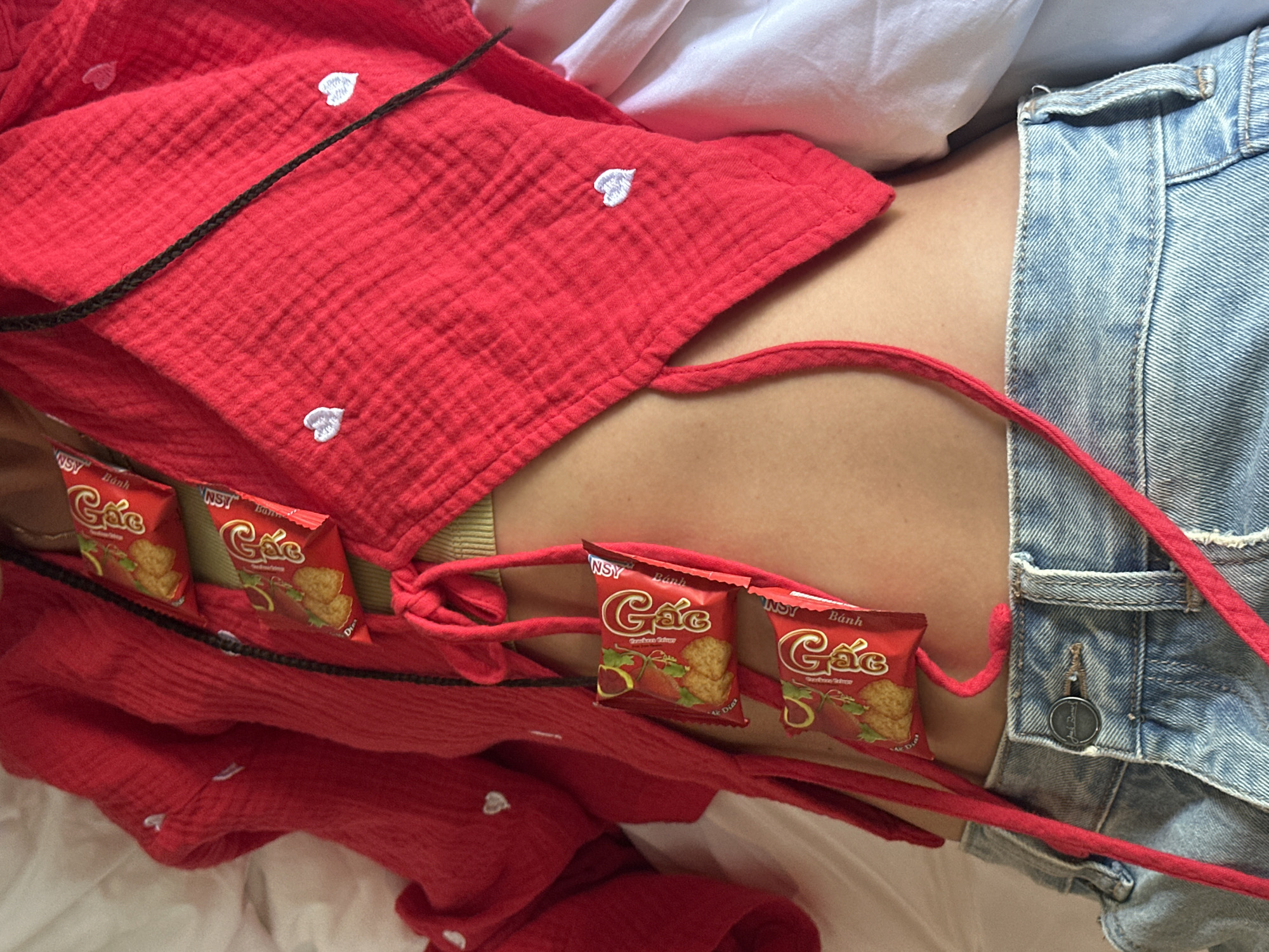 The image presents a close-up view of an individual's torso, from the chest to the upper thighs, in an indoor setting within Mù Cang Chải, Vietnam. The person is wearing a red, textured, long-sleeved top with small white embroidered heart patterns, featuring tie closures at the front. A light green ribbed tank top is visible beneath the red garment. Five small, red rectangular snack packages, each labeled "Gác Bánh" and "NST" with depictions of cookies and fruit, are strung together with red cords and draped vertically over the red top. A dark, braided string also runs down the center of the red top. The individual wears light blue denim jeans, partially unfastened, revealing a metal button embossed with "Jack & Jones." The background consists of white bedding material.