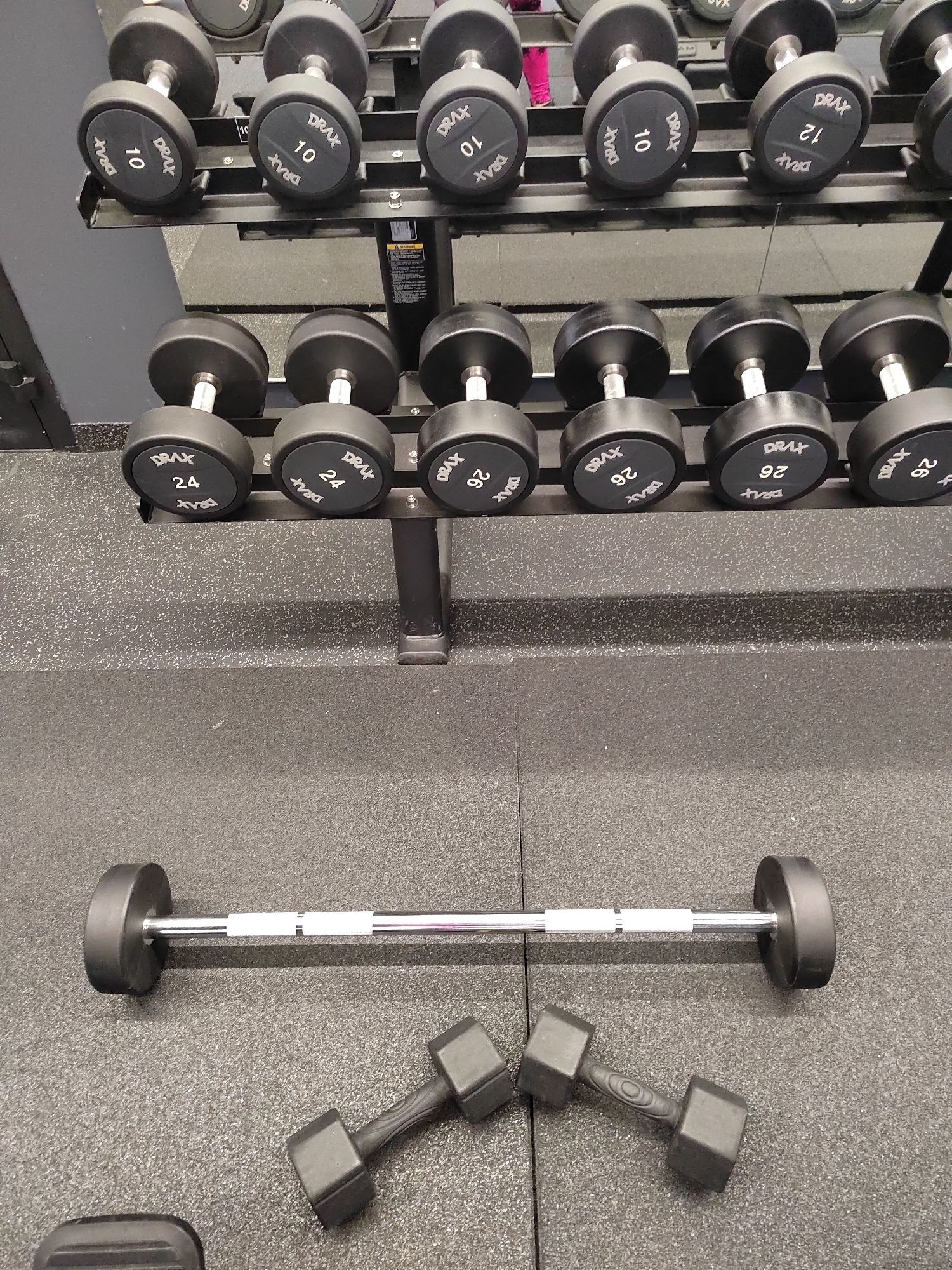 In a gym located in Novogorelovo, Russia, a variety of weightlifting equipment is displayed on racks and the floor. The upper rack holds dumbbells labeled with weights ranging from 10 to 12. The middle rack features dumbbells labeled 24 and 26. On the floor, a barbell with black end weights rests horizontally, accompanied by two smaller, black dumbbells with textured handles. The environment appears to be a clean, well-lit fitness facility, with dark grey rubber flooring and a dark grey wall in the background. There are no people visible in the image.