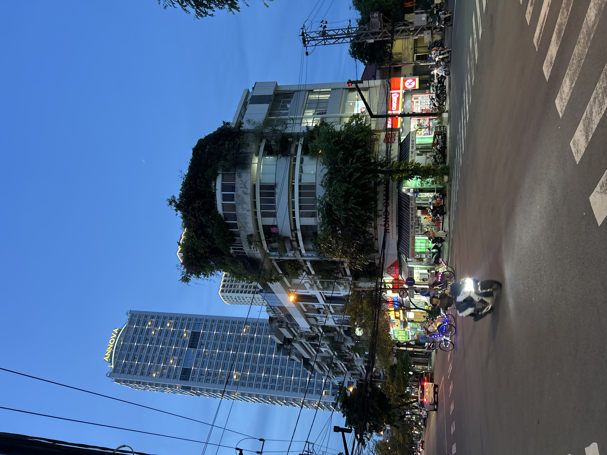 This photograph captures a vibrant street scene in Nha Trang Ward, Vietnam, at dusk or early evening. The sky, still a deep blue, suggests the transition from day to night, with artificial lights already illuminating the urban landscape.
The street is lined with a mix of buildings. On the left (when re-oriented correctly), a distinctive multi-story building features a curved facade heavily overgrown with lush green vines and foliage, creating a unique, organic appearance. Further back and to the left, a modern, tall skyscraper stands out with its illuminated sign spelling "ANNOVA" in yellow letters. Other commercial buildings with various light signs flank the street.
Activity on the street includes several motorbikes, a common mode of transport in Vietnam, parked along the sidewalk and a few moving on the road. One motorbike with a bright headlight is prominently visible in motion, and a white car can be seen further down. Several pedicabs or cyclos, some adorned with colorful lights, are also present. White zebra crossings are marked on the asphalt road. Overhead, a complex network of power lines and utility poles crisscrosses the sky, characteristic of many urban areas in the region.
Visible text on storefronts provides strong location cues. Signs for food establishments display "HỒNG GIANG" and "PHỞ + BÚN BÒ HUẾ," indicating popular Vietnamese noodle dishes. A "CIRCLE K" convenience store sign is also visible. The overall scene depicts a busy, typical evening in an Asian city, with a blend of modern architecture and traditional street life.