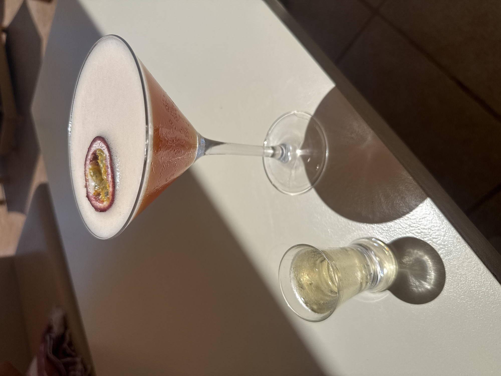 An outdoor close-up shot shows a passion fruit martini and a shot glass filled with a pale yellow liquid on a pale gray surface. The martini, served in a martini glass, has a frothy pinkish-orange liquid topped with a slice of passion fruit. The shot glass, situated to the left of the martini glass, contains a clear, bubbly liquid. Shadows are cast on the surface from both glasses, indicating sunlight. In the background, out of focus, is a hint of a folded purple cloth on a lighter surface, possibly a lounge chair. The scene appears to be set in an outdoor seating area, possibly at a bar or restaurant, in the city of Llucmajor, Spain, given the bright sunlight and outdoor setting.