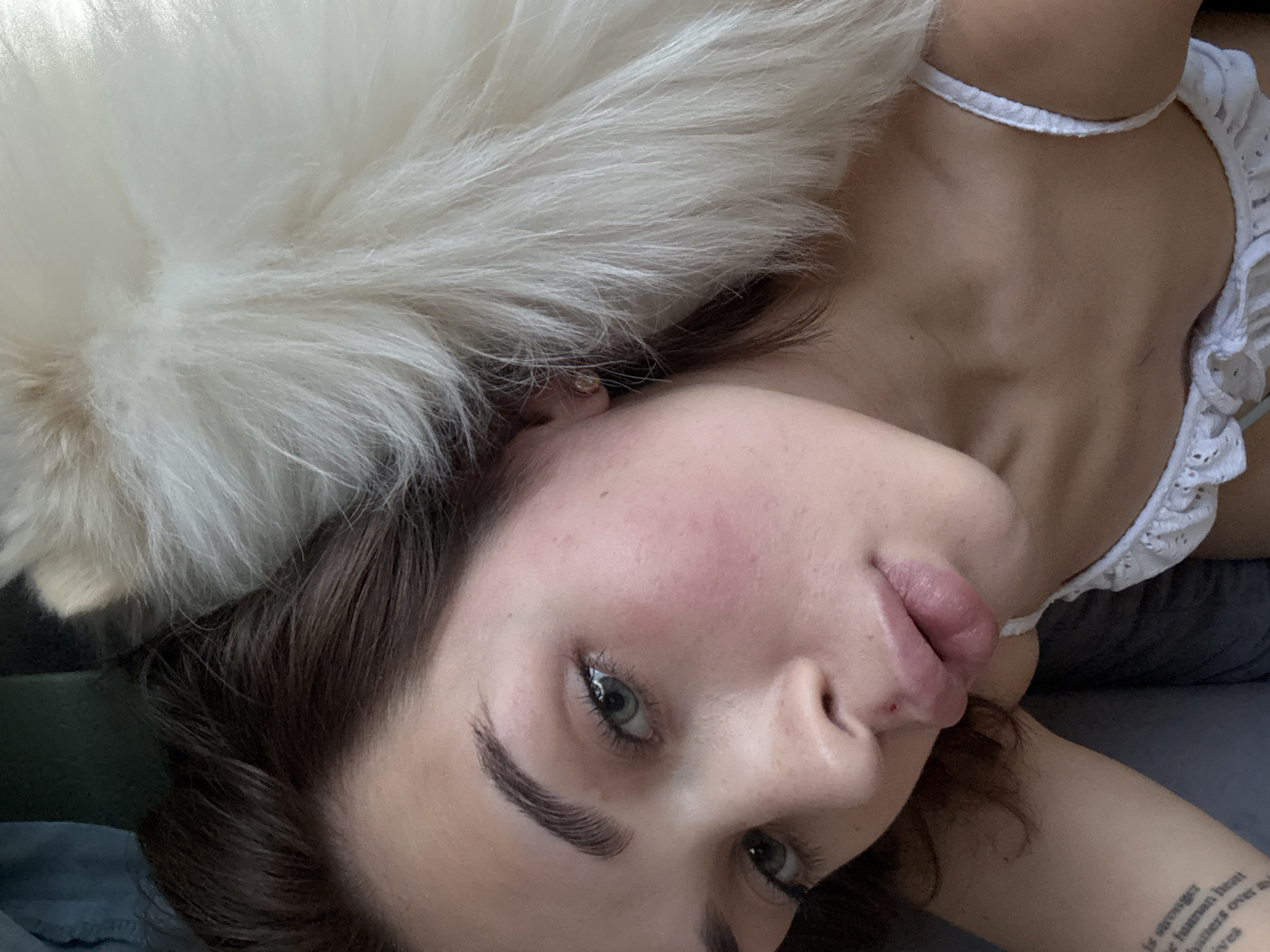 The image depicts a woman lying down, looking at the camera, with a white fluffy dog positioned near her head. She has brown hair, fair skin, and is wearing a white garment with lace. Part of a tattoo is visible on her right arm. The background is indistinct, with a neutral gray tone. The location in the city of Лобаново, Russia, cannot be determined from the visual information provided.