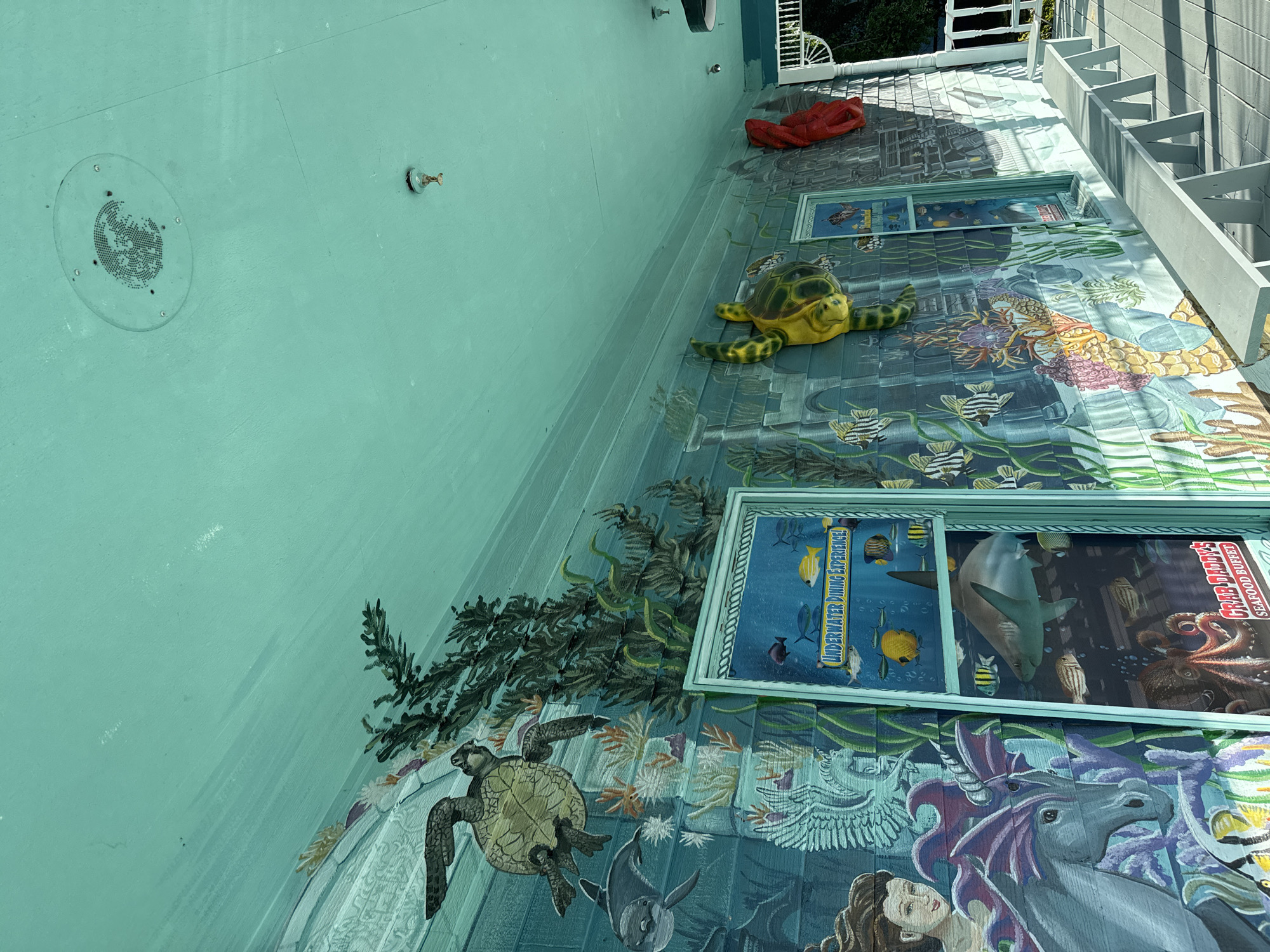 The image shows a building exterior in Garden City Beach, United States of America, decorated with an underwater-themed mural. The mural features painted sea creatures like turtles, fish, and a shark. Three-dimensional sculptures of a turtle and a lobster adorn the mural. Framed signs read "UNDERWATER DINING EXPERIENCE!" and "GREEDY PIRATE'S SEAFOOD BUFFET" are present. A mermaid and a unicorn are also painted on the bottom section of the mural. The building is painted a light turquoise color. A set of exterior stairs is visible.
