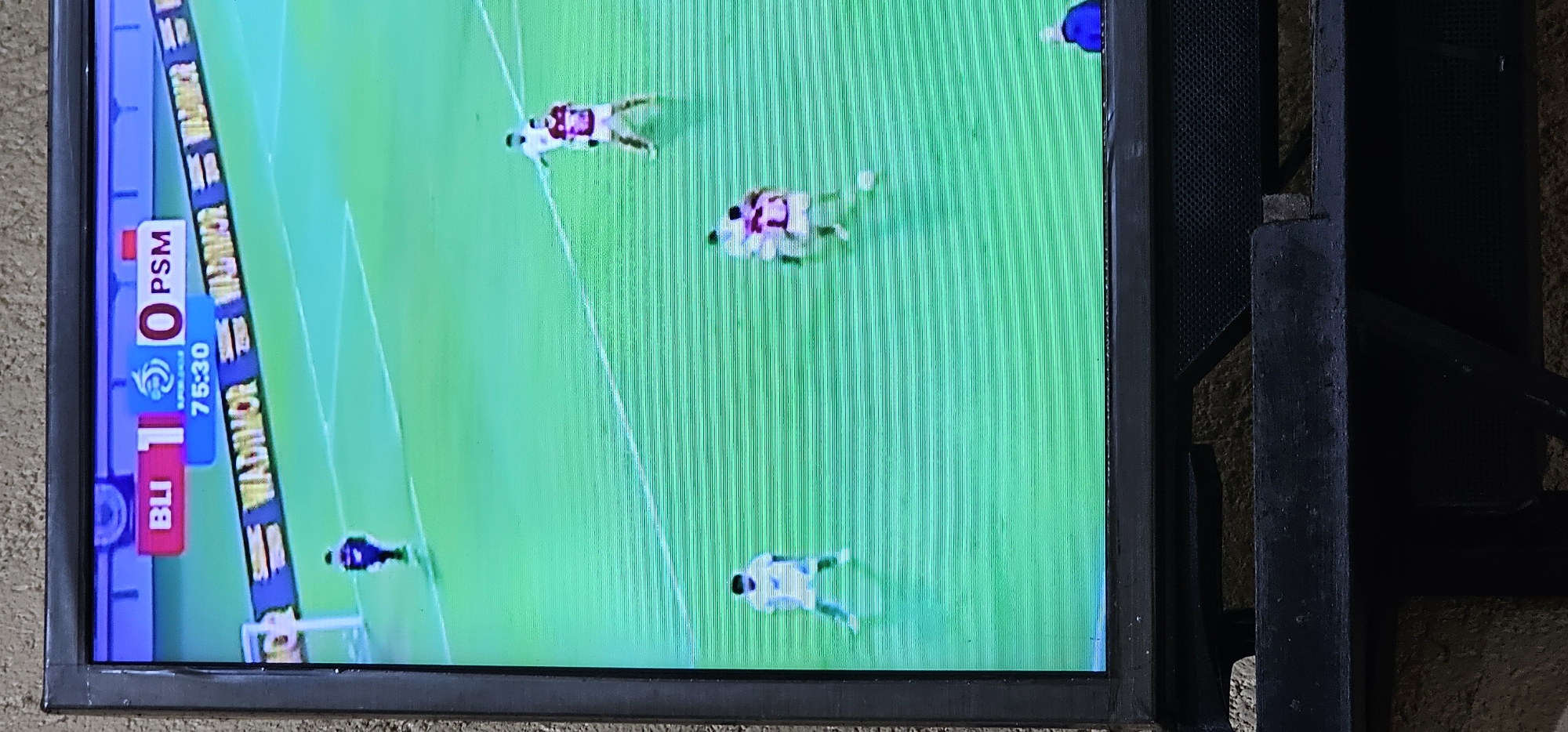 The image shows a TV screen displaying a soccer match between Bali United (BLI) and PSM. The score is 1-0 in favor of Bali United, and the game clock shows 75:30. The location on the screen mentions "Wadmor". The game is being played on a green field with white lines. Several soccer players are visible on the field. The TV is in an indoor setting, next to a dark, metallic-looking structure. The TV appears to be resting on a wall or surface. Based on the teams, the scene is likely in Denpasar, Indonesia.