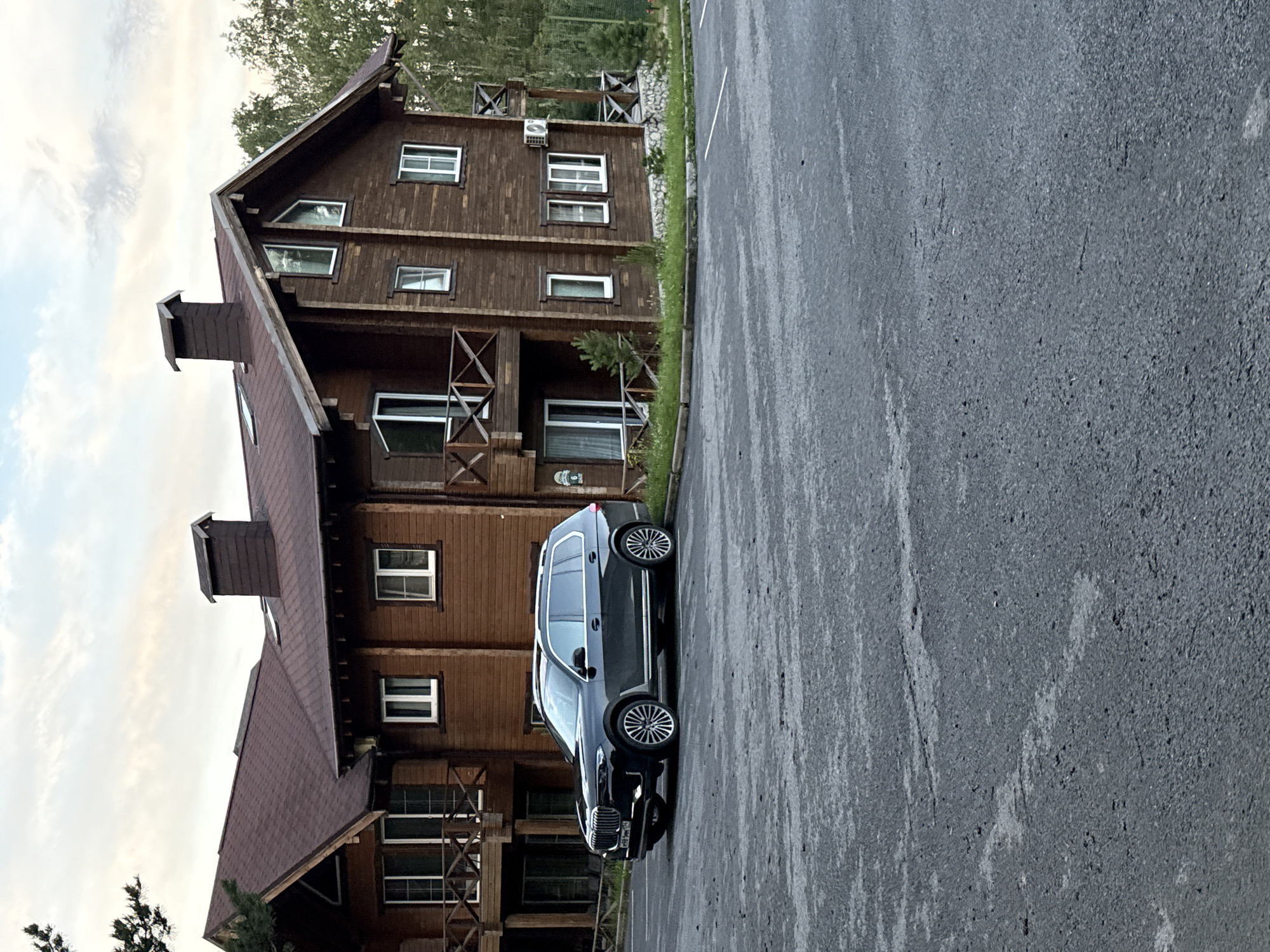 A dark-colored luxury SUV is parked on a wet asphalt surface in front of a large, multi-story wooden building. The building features a dark red shingled roof with visible chimneys and wooden balconies. The facade of the building is constructed of dark wooden planks, with numerous windows of varying sizes, some with white frames. There is a patch of green grass and some small pine trees visible between the parking lot and the building. The background shows trees and a cloudy sky. The image appears to be taken in Syrostan, Russia.