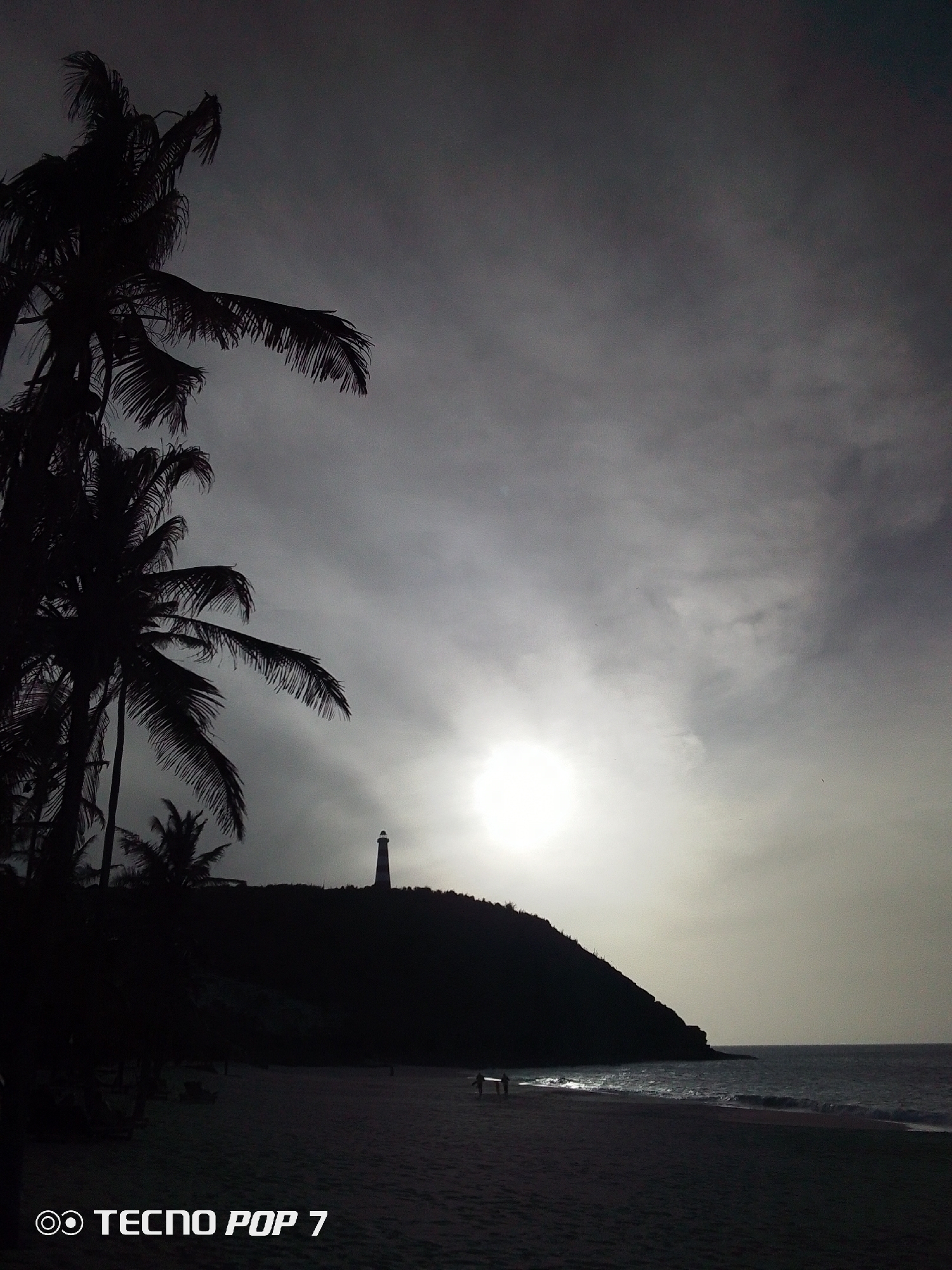 The image depicts a coastal scene in Pedro González, Venezuela, dominated by a dark silhouette of a promontory topped with a lighthouse against a bright, overcast sky. The beach in the foreground is dark, with visible sand texture and faint suggestions of small figures walking along the shoreline. Palm trees are silhouetted in the left portion of the frame, and the ocean extends into the distance. The sun is positioned behind the hill, creating a hazy glow around the lighthouse. The lower left corner displays the text "TECNO POP 7", indicating the device used to capture the image.