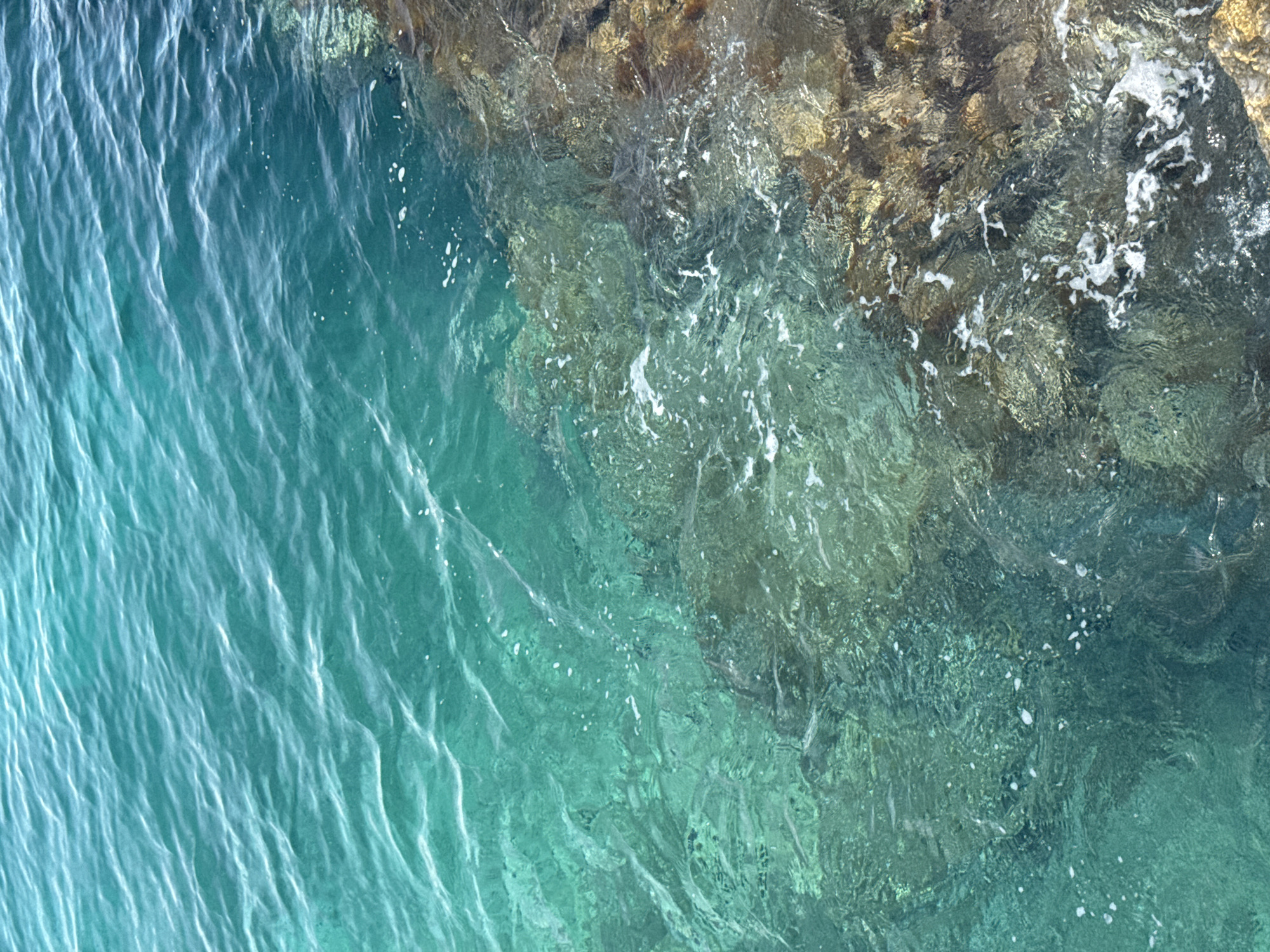 This media file is an aerial shot of the Adriatic Sea at Dubraljevina, Montenegro. The image captures the transition between the deep turquoise waters of the open sea and the shallow, clear water near a rocky shore. Sunlight glistens on the surface, creating dynamic patterns of light and shadow on the rippling water. The rocky edge of the coastline is visible in the upper right quadrant of the image, with a mixture of brown and grey rock formations. The water near the shore is exceptionally clear, revealing the submerged rocks and seabed beneath. Small white foam bubbles are scattered across the water, indicating gentle wave action against the rocks. The overall impression is one of natural beauty and tranquility, with the vibrant colors of the sea and the ruggedness of the coastline creating a picturesque scene. The time of day appears to be daytime, likely late morning or early afternoon, given the bright sunlight. No people or man-made objects are visible.