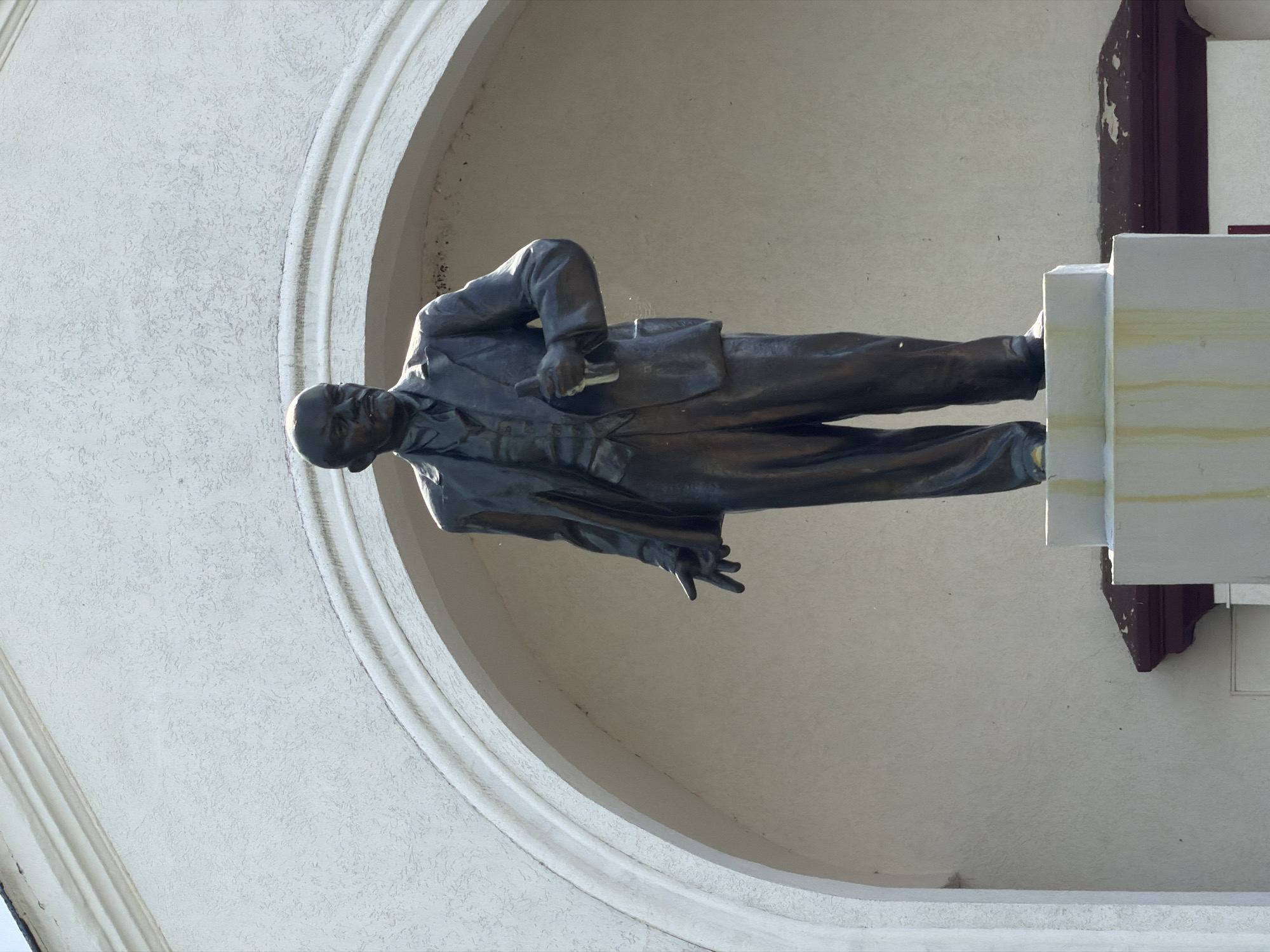 A bronze statue of a man dressed in a suit and tie stands within a building's archway. The statue is oriented horizontally, with the man's head on the left and his feet on the right. He holds a rectangular object in his left hand and his right hand is extended forward with his fingers splayed. The background consists of a textured, light-colored wall and a curved architectural element. To the right of the statue, a portion of a pedestal and a dark architectural trim are visible. The lighting suggests it is daytime, with shadows indicating the sun is shining. The statue appears to be located in a public space, possibly a square or memorial, given its monument-like presentation. The location context provided is Бягомльскі сельскі Савет, Belarus, suggesting this is a landmark within that region. There is no visible text in the image. The overall mood is solemn and commemorative.