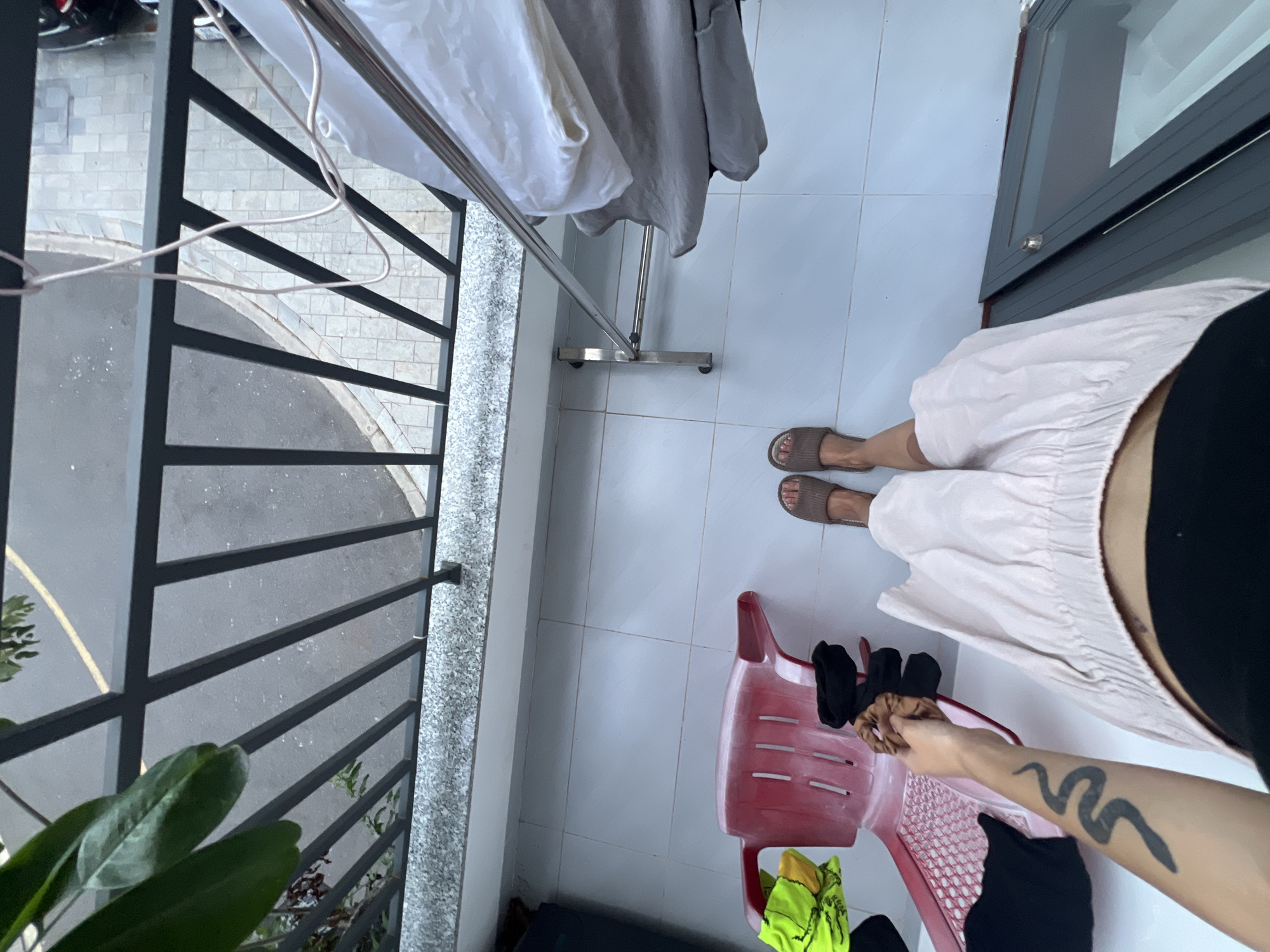 Captured from an elevated, head-on perspective, the image presents a partial view of a balcony in Nam Nha Trang Ward, Vietnam. A person wearing brown sandals and a long, light-colored skirt, partially obstructed, stands on the white-tiled balcony. The person's arm, featuring a tattoo of a snake, is visible. A red plastic chair sits on the balcony, with some clothing items. A clothing rack with hanging clothes stands near the center of the frame. The balcony is enclosed by a dark metal railing with a view of the street. Plants are seen at the lower-left corner of the frame. A sliding glass door is visible on the right. The environment appears to be residential.