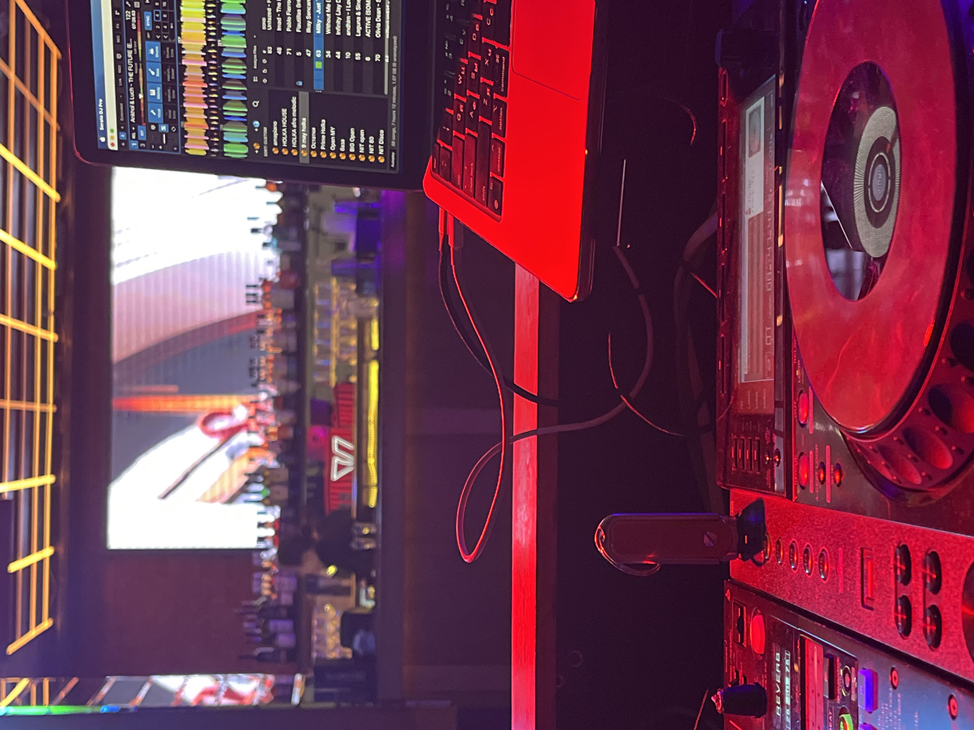 The image shows a DJ setup, including a laptop displaying Serato DJ Pro software, a DJ controller, and a USB drive. The laptop screen displays a music playlist with song titles such as "Ankhol & Luch - THE FUTURE (E...)," "HOLKA HOUSE," and "9 may holka." The DJ controller has various buttons and dials. The environment is dimly lit, with a bar in the background filled with bottles. The bar's neon sign is partially visible.