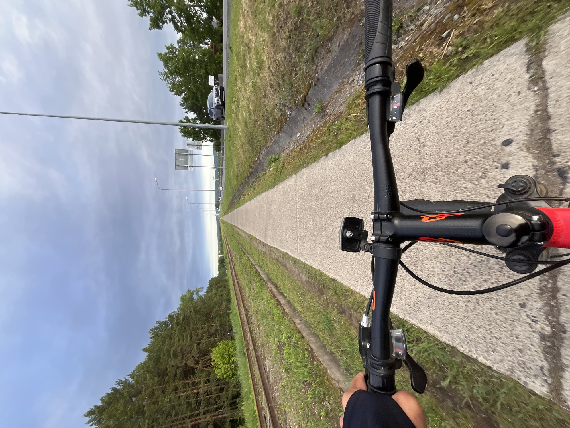 A first-person perspective photo shows a person riding a bicycle on a paved path. The path runs parallel to abandoned train tracks overgrown with grass. On the other side of the path, there is a road with trees and street lights. The sky is overcast. The location appears to be in Chaikovsky, Russia.