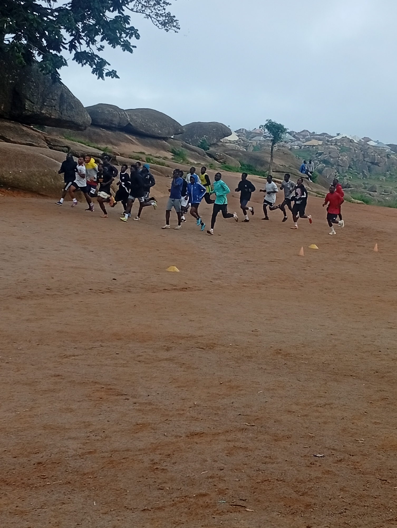 In Jos, Nigeria, a group of young men is engaged in a cross-country race on a dirt track. The race is taking place in a rocky, uneven terrain, with large boulders and sparse vegetation in the background. The sky is overcast, suggesting a cloudy day. The runners are wearing various athletic attire, including t-shirts, shorts, and running shoes. Some are in motion, actively running, while others are in the process of starting or finishing the race. The scene captures a moment of athletic activity amidst a natural, rugged landscape.