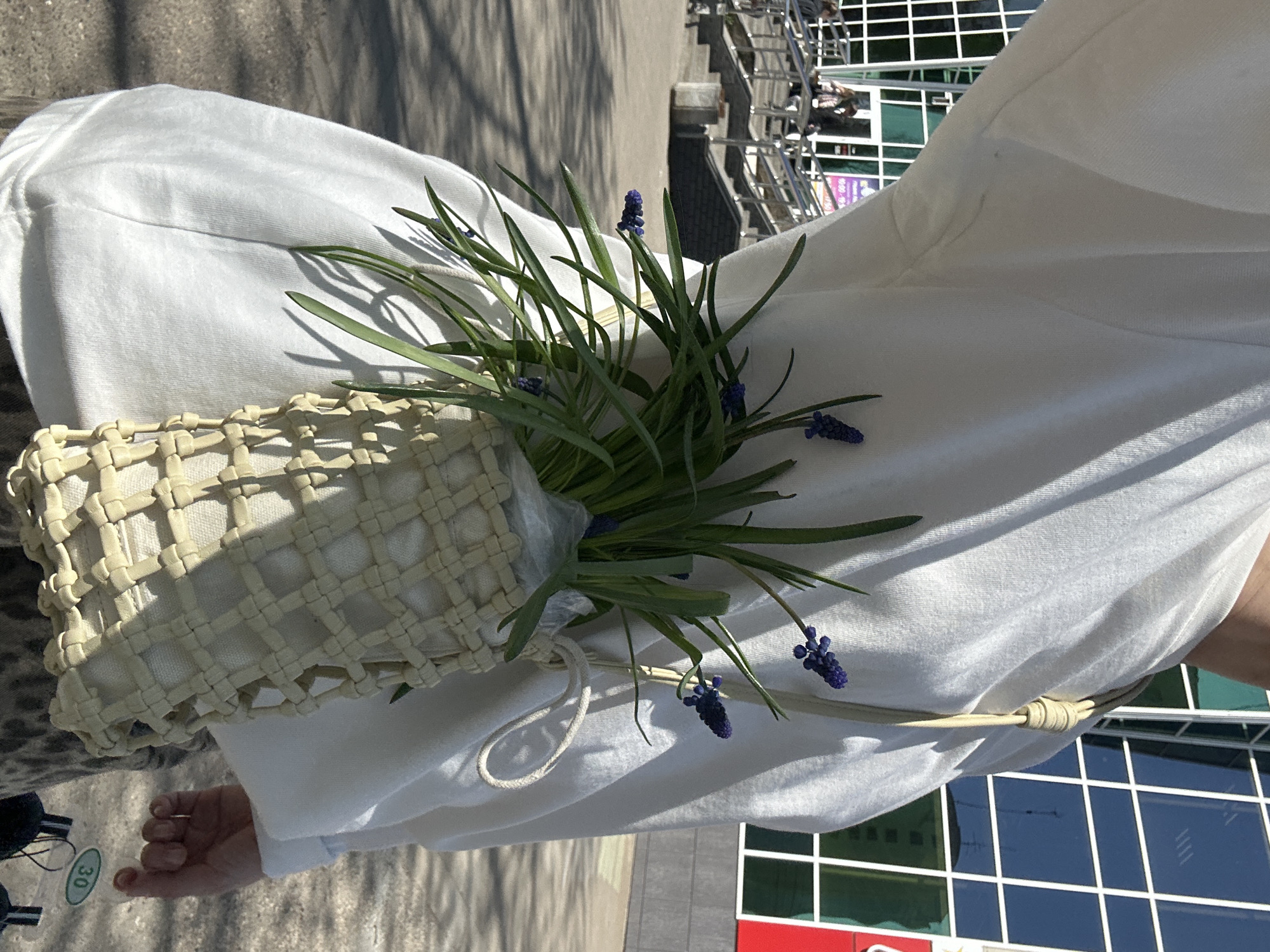 A person holds a woven bag containing grape hyacinth flowers. The person wears a white garment. The photograph appears to have been taken in an urban environment, possibly near the city of Chaikovsky, Russia, based on the location tags. The background shows city buildings and street infrastructure.