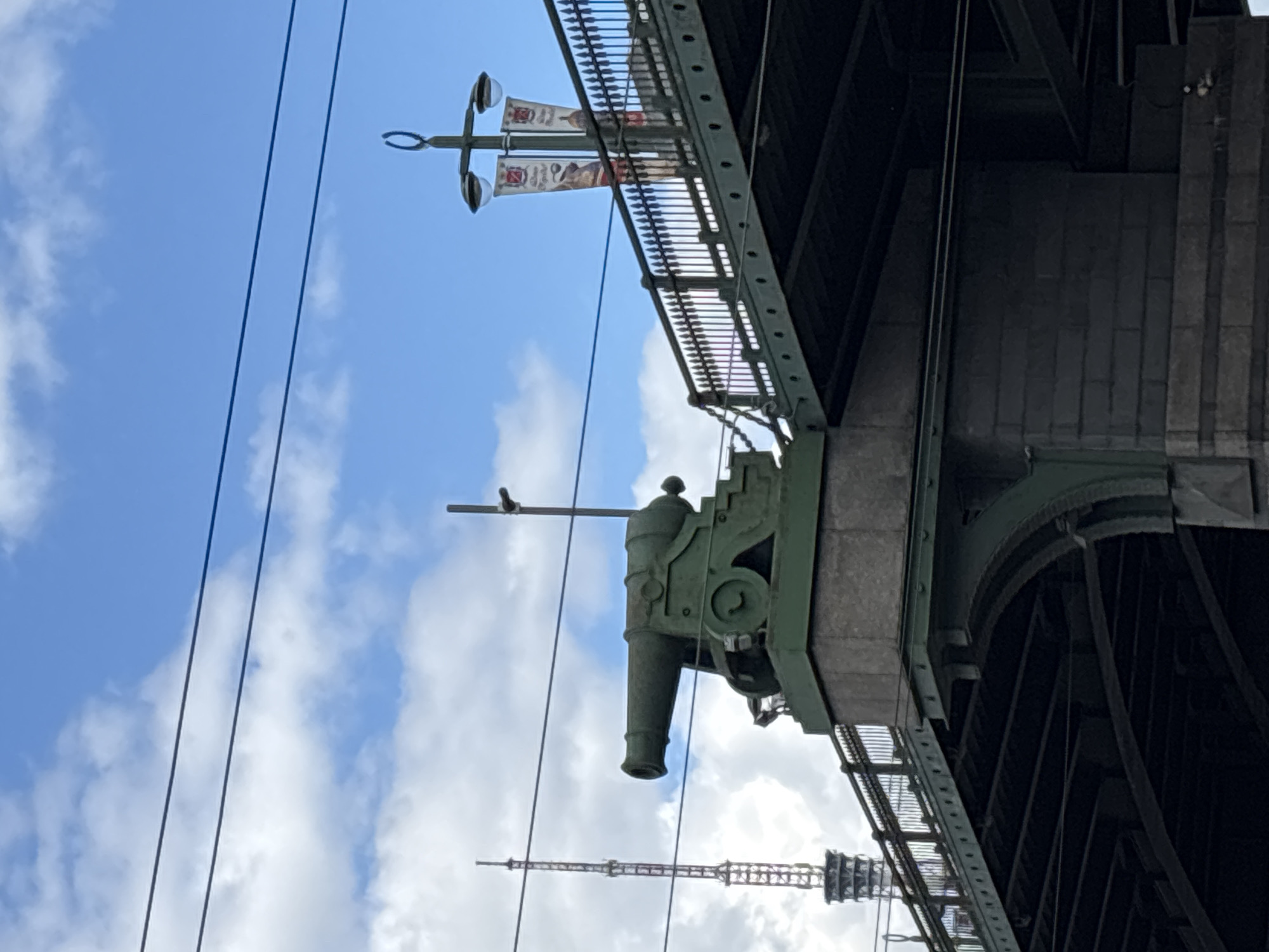 View is of a section of the Liteyny Bridge in Saint Petersburg, Russia. The bridge section is green and includes a decorative cannon mounted at an angle. Power lines are visible in the lower left and center of the frame. The sky is light blue with white clouds. In the distance is a tower.