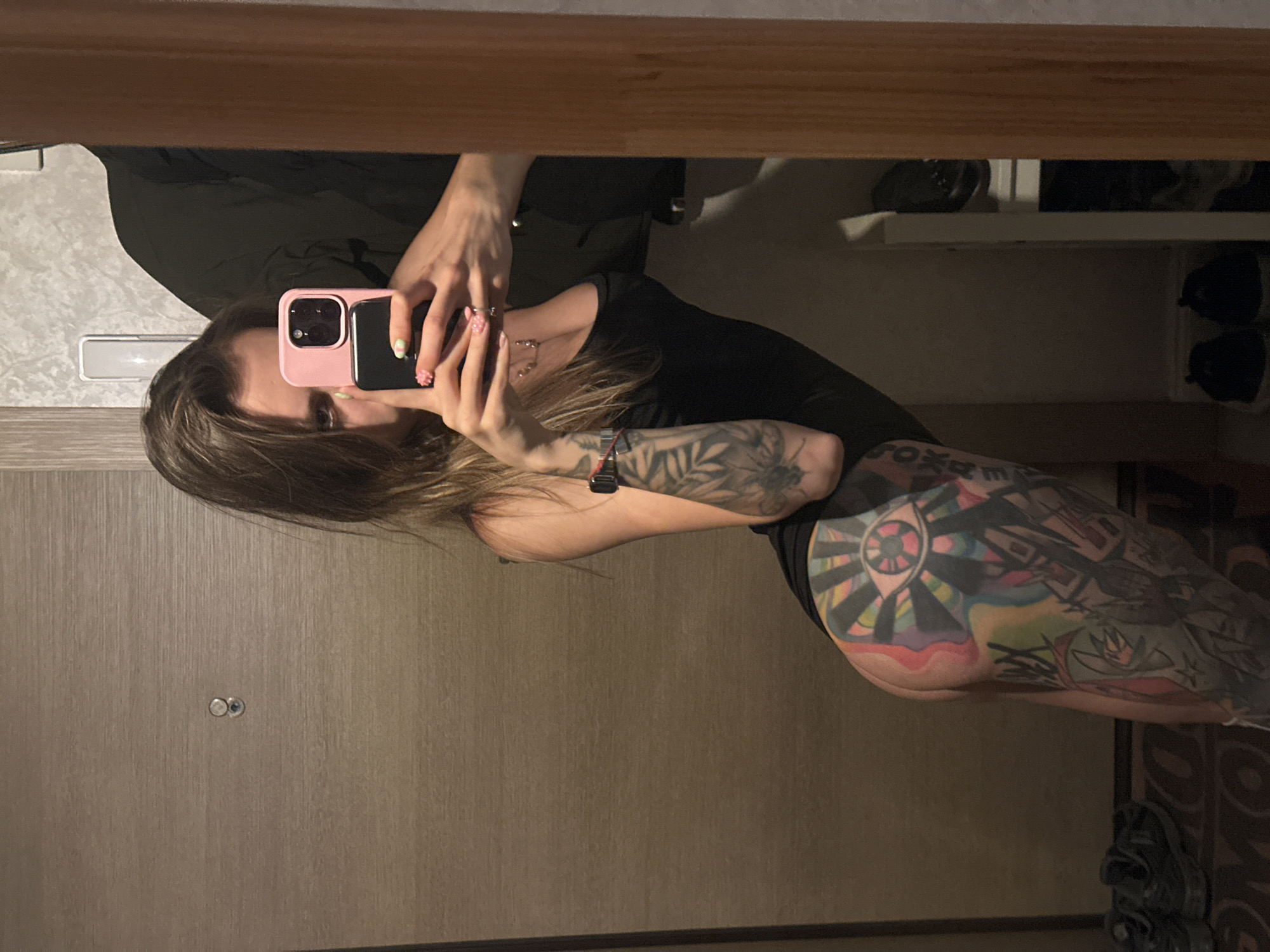 A woman is taking a selfie in a mirror. She has long, brown hair, and is wearing a black top. She has tattoos on her arm and leg. The room appears to be a hallway, with a wooden door and a wooden frame visible. There are shelves with shoes in the background. The lighting suggests it is daytime.