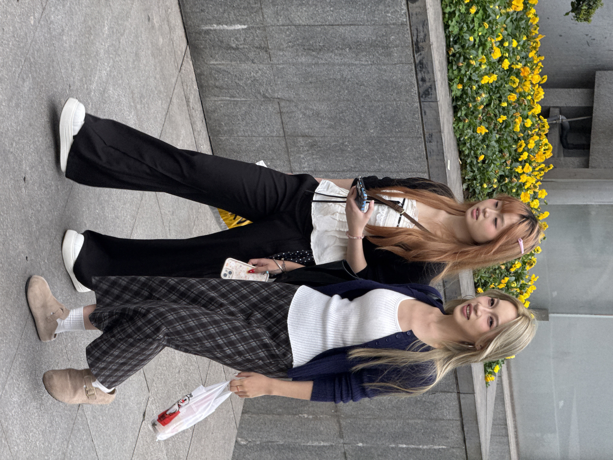 Two young women stand on a gray concrete sidewalk near a low wall and a border of yellow flowers, likely in Liangjiang, China. The woman on top has light brown hair and is holding a smartphone in her right hand and a white smartphone in her left hand. She is wearing white sneakers, black pants, a black jacket, and a white tank top. The other woman, closer to the viewer, has blonde hair and is holding a white plastic shopping bag in her left hand. She is wearing light brown shoes, a white tank top, a navy blue cardigan, and plaid pants.