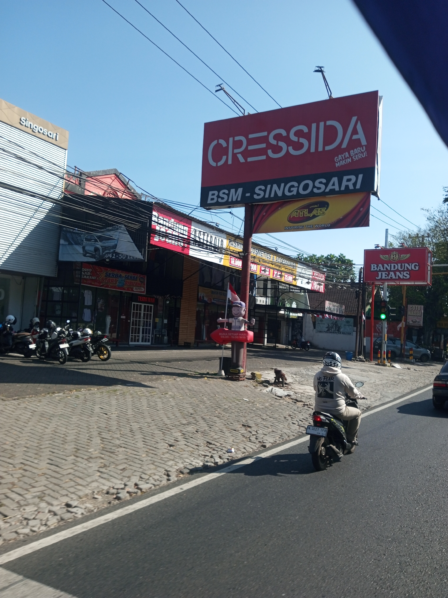 The image depicts a street scene in Ardimulyo, Indonesia, on a clear, sunny day. A street lined with various shops and billboards runs horizontally across the frame. On the left, a building with a modern, metallic facade is branded "Singosari". Adjacent to it, a red-tiled building features signs for "Cressiida" and "BSM-Singosari". Further down the street, more shops with signs like "Atlas" and "Bandung Jeans" are visible. A traffic light is positioned on the right side of the road, showing a green light. Several motorcycles are parked along the sidewalk in front of the shops, and one motorcyclist, wearing a light-colored hoodie with "NO FEAR" printed on the back, is riding away from the camera in the right lane of the road. The pavement consists of interlocking grey bricks, and the road surface is asphalt with a white line marking the lane. The environment appears to be a commercial area with pedestrian and vehicular traffic.