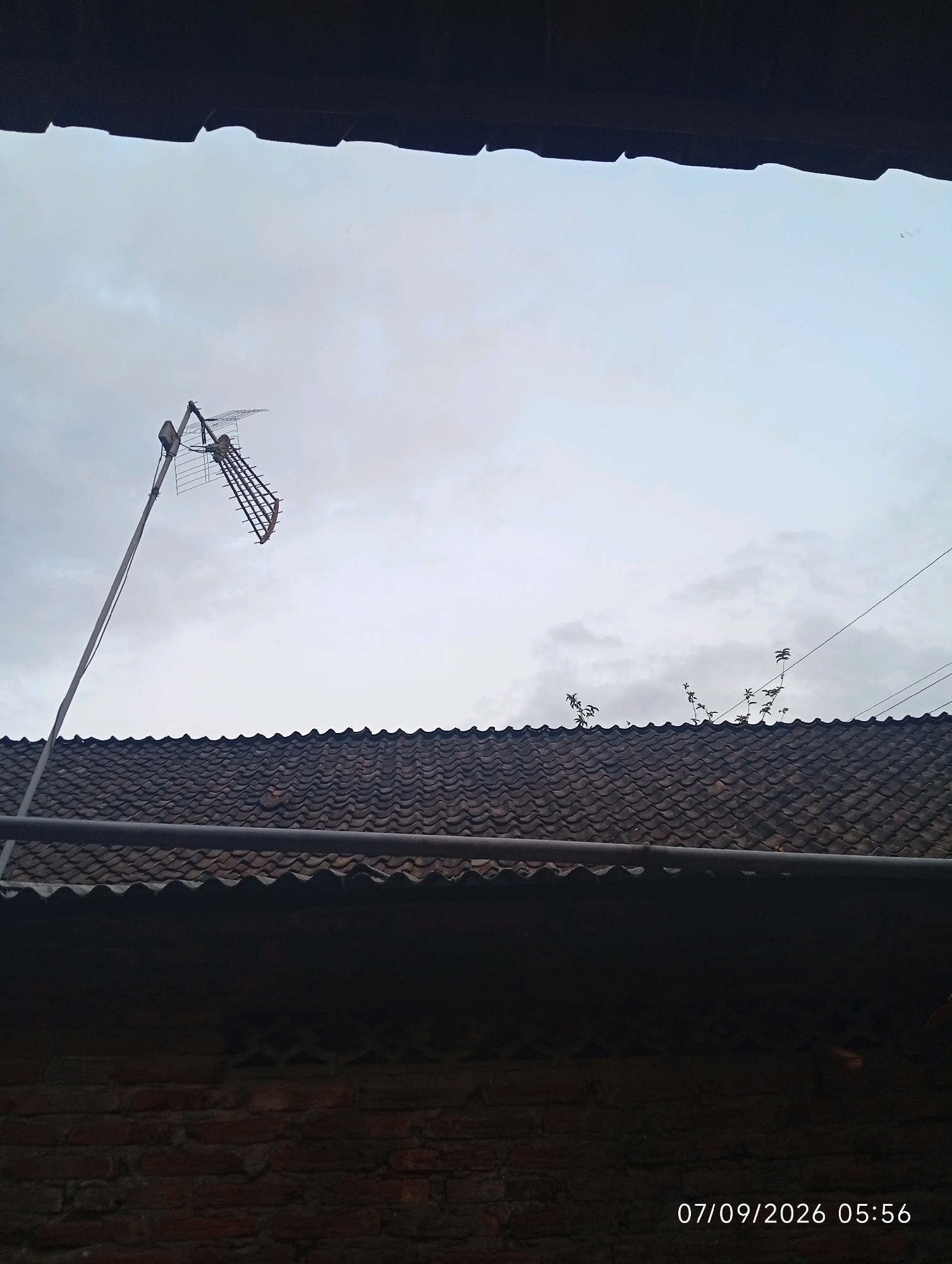 The image shows an outdoor scene with a brick wall in the foreground, a tiled roof, and a television antenna. The sky is cloudy and overcast. A date and time stamp, "07/09/2026 05:56," is visible in the lower right corner. There are no people or specific identifiable landmarks in the image that would indicate the exact location in Gebang, Indonesia, beyond it being an outdoor residential or urban setting.