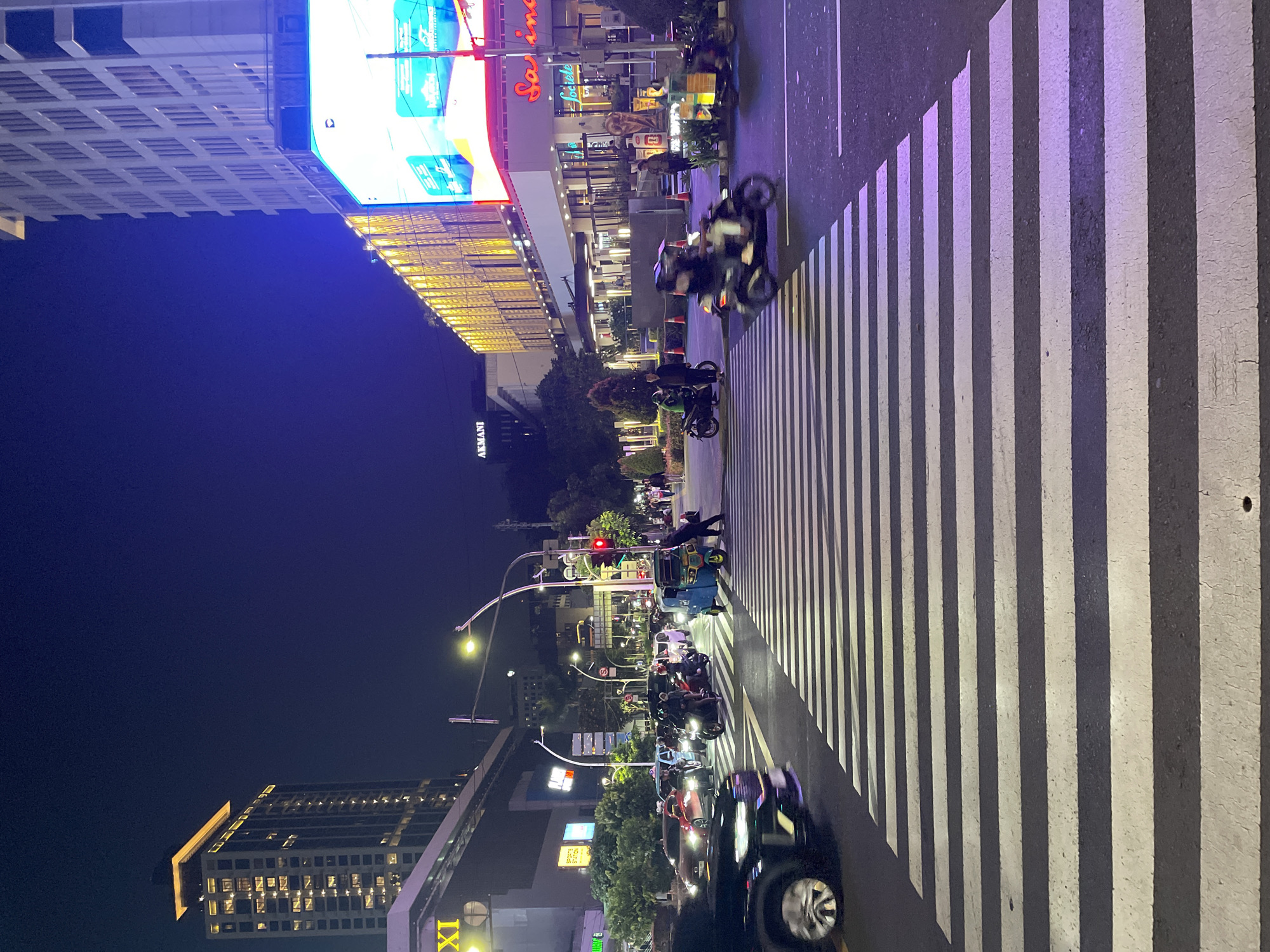 The image depicts a night scene in Central Jakarta, Indonesia, featuring a crosswalk illuminated by artificial light. Several motorcycles and cars are visible on the street, some in motion. Pedestrians can be seen crossing the street using the crosswalk. A tall building with an illuminated billboard stands to the left of the street. The environment is urban, with streetlights and commercial buildings lining the road. The sky is dark.