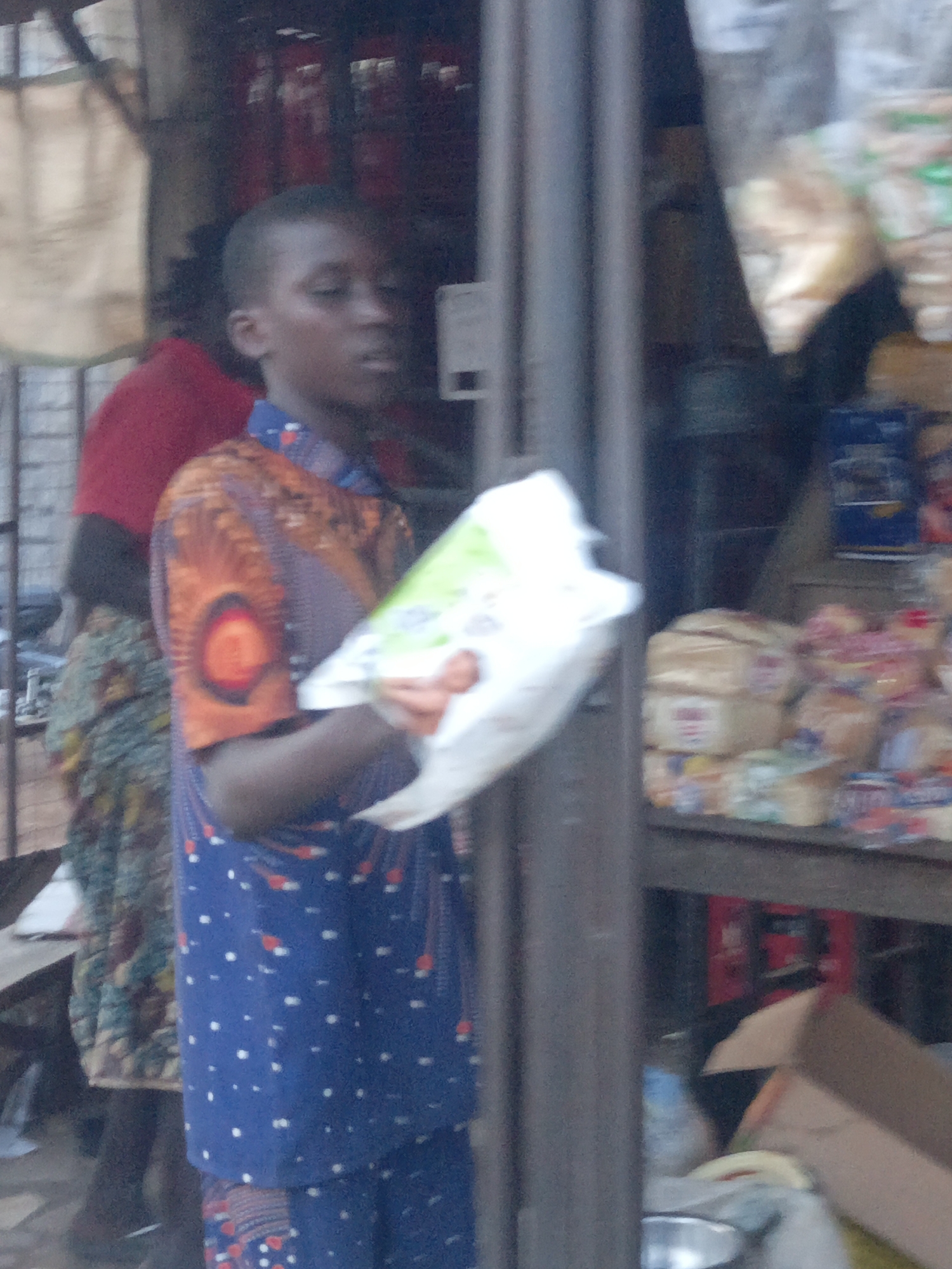 A person stands in front of a shop in Akwanga, Nigeria, holding a white plastic bag. The shop displays items like bread and snacks. Other people and objects such as metal bars, a cardboard box, and beverage crates are also present.