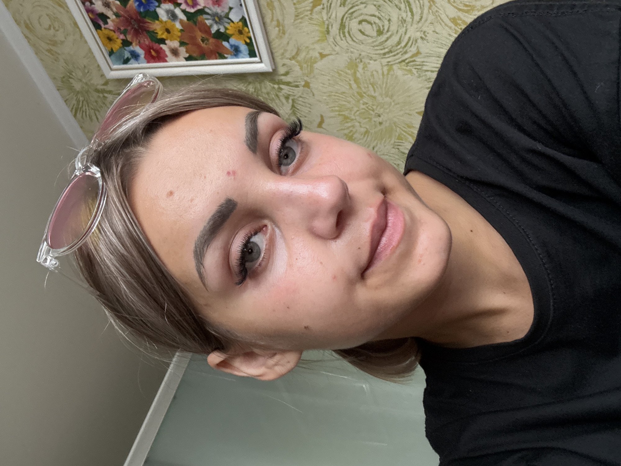 A young woman with blonde hair tied in a ponytail is looking at the camera. She is wearing a black t-shirt and pink sunglasses are perched on her head. The background shows a wall with floral wallpaper and a framed painting of flowers. The scene appears to be indoors, possibly in a residential setting.