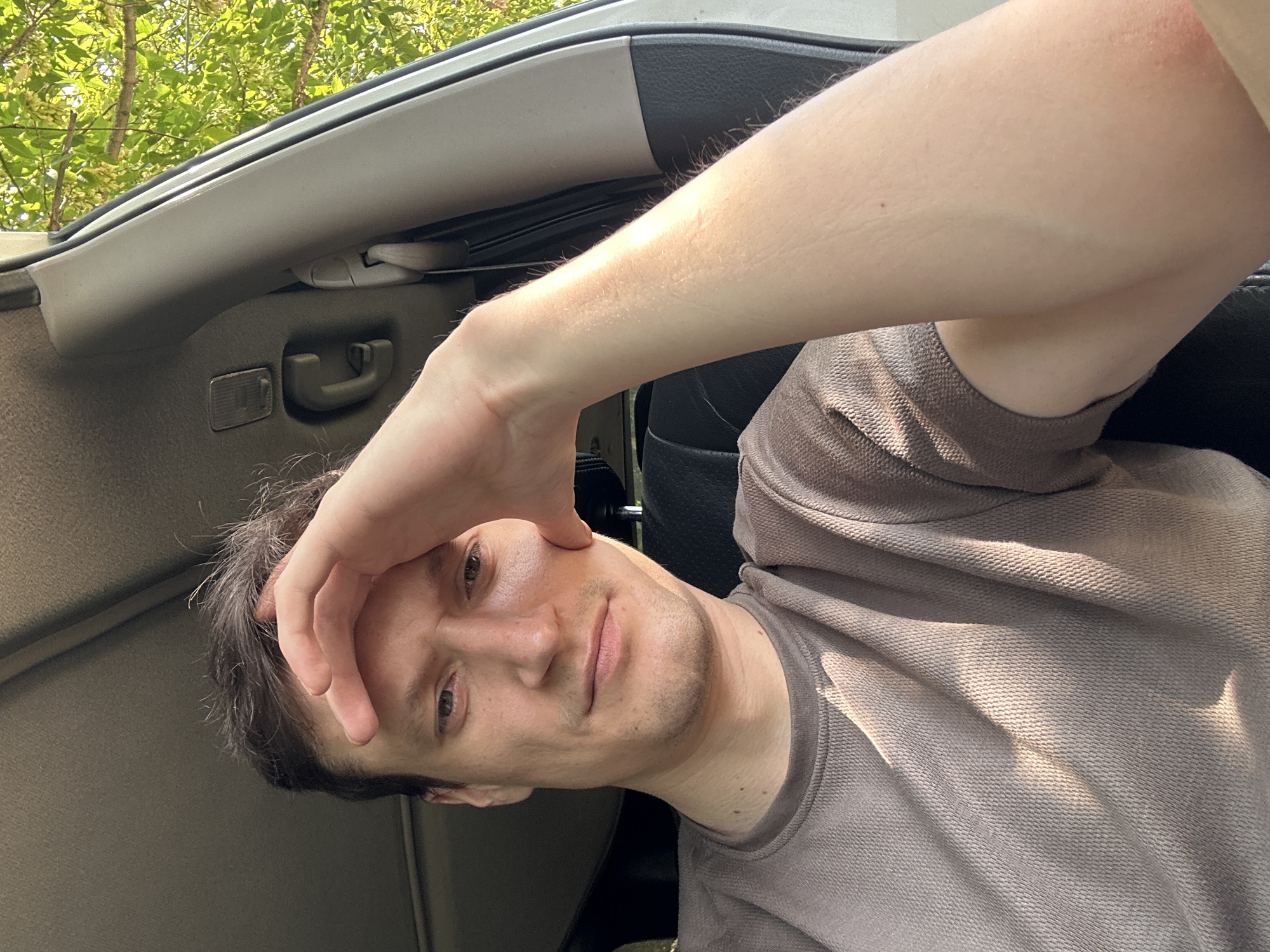 In the image, a man is seen from the chest up, seated inside a vehicle. He has his right hand resting on his forehead, and his left arm is bent at the elbow with his hand on his cheek. The man is wearing a grey, long-sleeved t-shirt. Sunlight is casting shadows on his face and t-shirt. Outside the vehicle, green foliage is visible, suggesting a location with trees. The interior of the car shows part of the car door and a headrest. The scene appears to be captured during daylight. There is no information in the image to indicate the specific location in the city of Makiv, Ukraine.