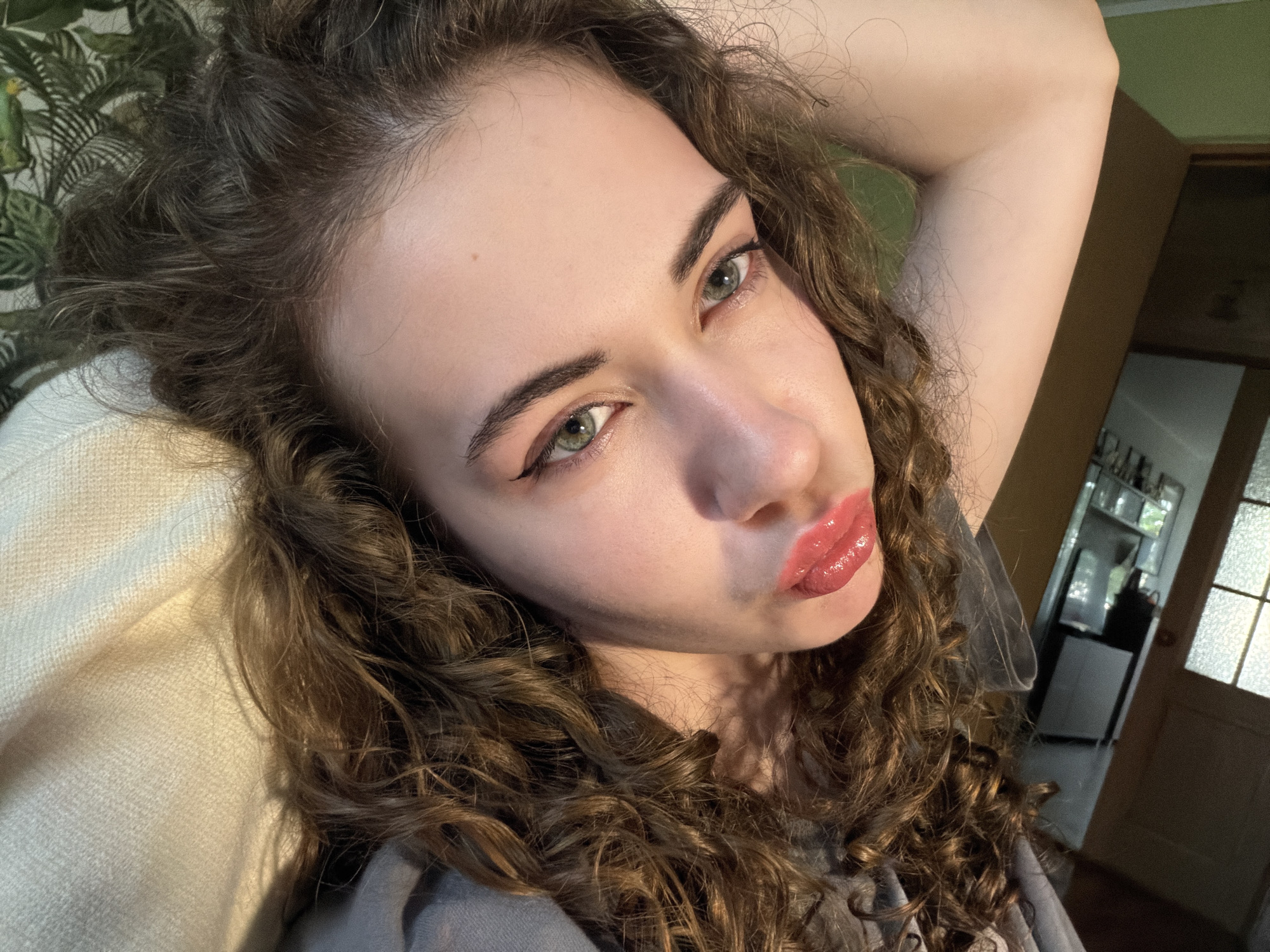The media file is a close-up, low-angle selfie of a young woman with long, curly brown hair. She is lying down, with her head resting on a cream-colored textured surface, possibly a blanket or cushion. Her left arm is raised and bent behind her head, and her right hand is not visible.
The woman has fair skin, prominent eyebrows, and striking light green eyes. She is wearing makeup, including eyeliner that creates a winged effect and glossy pink lipstick, giving her lips a plump appearance. She is looking directly at the camera with a slightly puckered expression.
The background is partially visible, revealing a wall with a green and white tropical-patterned wallpaper on the left. To the right, there is a glimpse of an interior space with a doorway and shelves, suggesting a home environment. The lighting suggests it is daytime, with natural light casting shadows.
The overall mood is casual and intimate, with the subject appearing relaxed and confident. There is no visible text in the image. The provided location context is Zavodoukovsk, Russia.