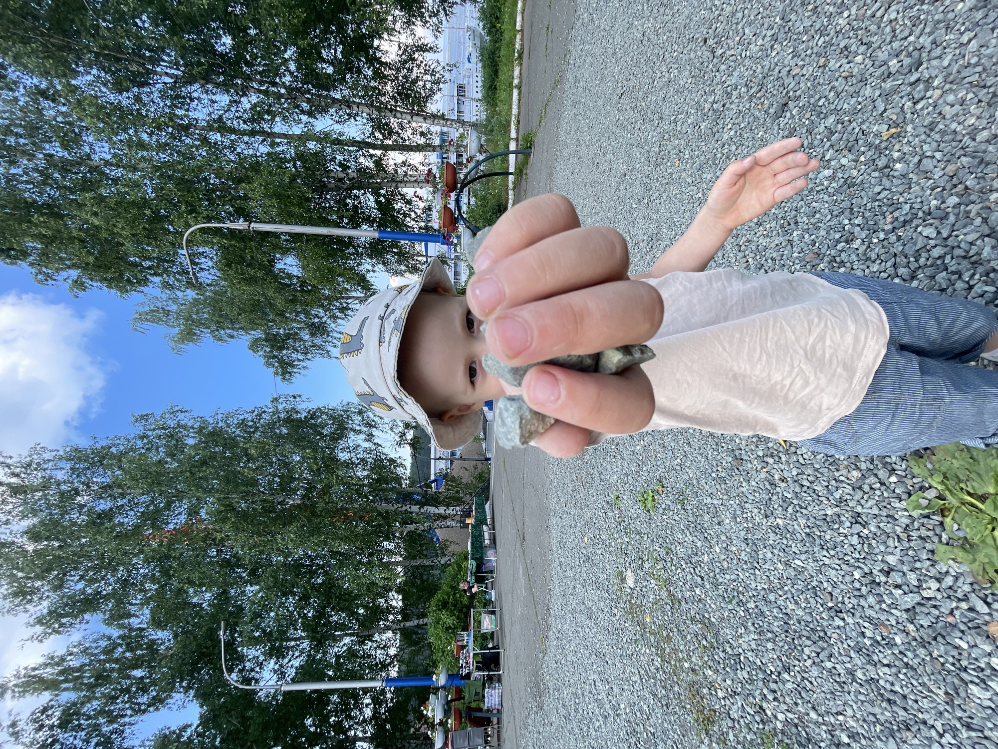 In the city of Chaikovsky, Russia, a young child wearing a bucket hat and a light-colored tank top over blue striped pants is holding a handful of small gray stones towards the camera. The child's face is partially visible, with direct eye contact. The ground is covered in small gray gravel, and tall birch trees with green foliage line the background. In the distance, there are some market stalls and a large white boat on a body of water. The sky is blue with scattered clouds.