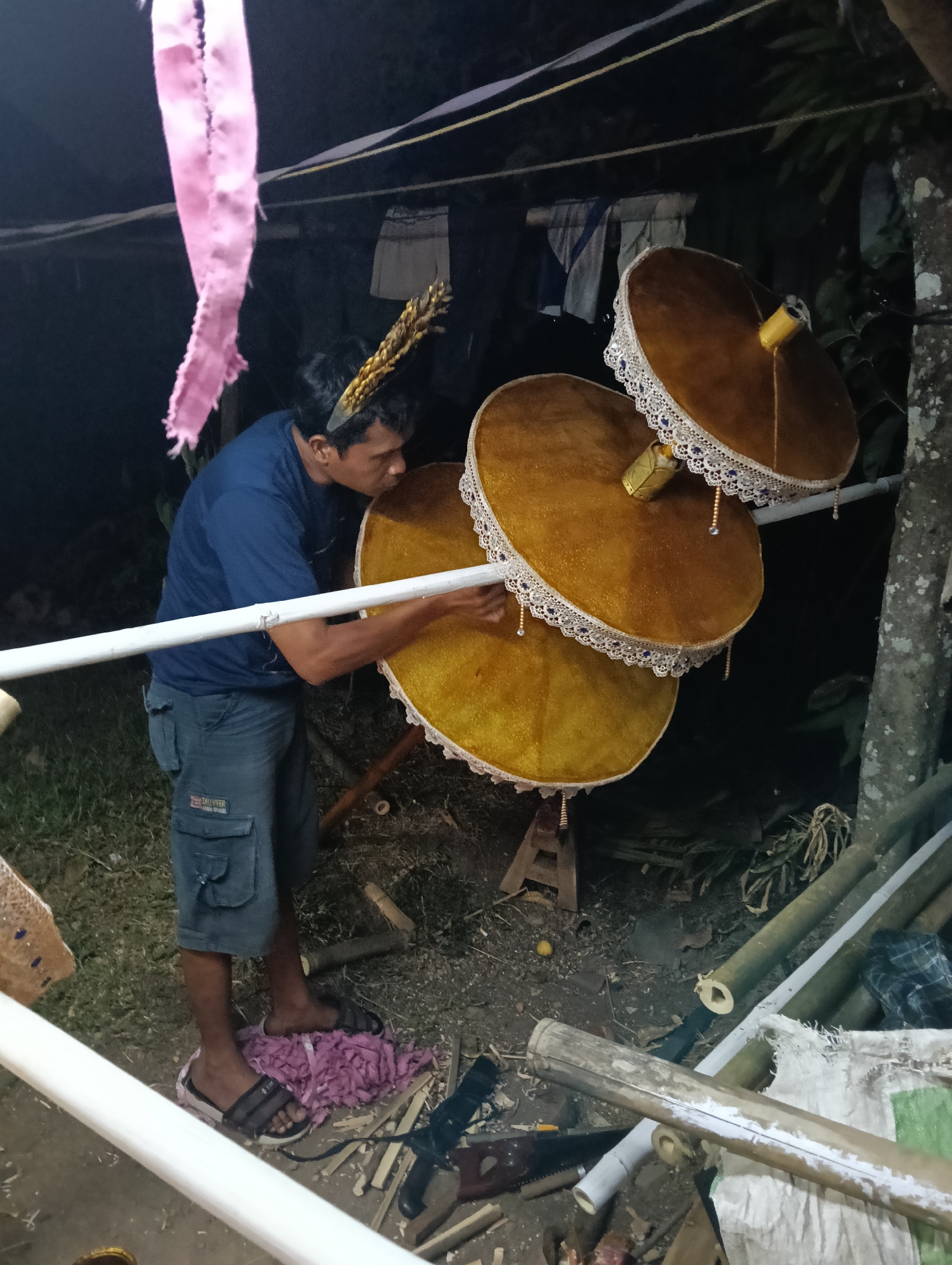 A man in a blue t-shirt and denim shorts is working on a series of three golden, circular parasols, stacked vertically and adorned with white lace trim. He is in an outdoor setting, possibly a yard, with a dark, natural background suggesting nighttime or a dimly lit area. The ground is covered with dirt, grass, and what appears to be wood scraps, indicating a work in progress. Several lengths of bamboo and other construction materials are visible. The man is actively adjusting one of the parasols, his hands engaged with the assembly. A pink fabric streamer hangs from above to the left, and a tree trunk is visible on the right side of the frame. The overall impression is one of a craftsman at work on decorative items, likely for an event or ceremony. Based on the stylistic elements of the parasols and the surrounding environment, the scene is consistent with cultural practices found in Tasikmalaya, Indonesia, where such ornate umbrellas are often used in local traditions.