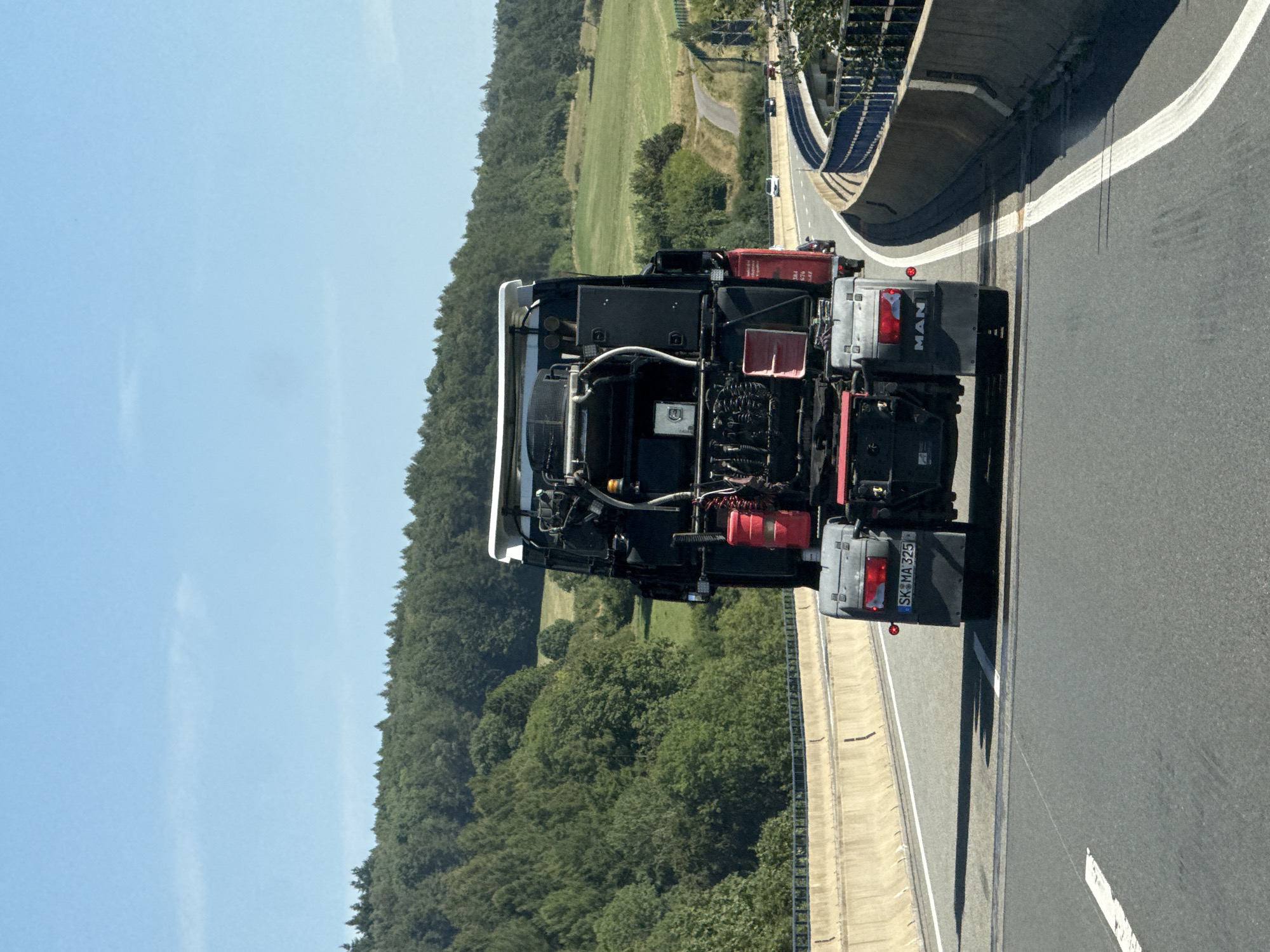 An overhead shot captures the rear of a dark-colored MAN semi-trailer truck driving on a highway. The truck is equipped with a red fire extinguisher mounted on its side. The license plate reads "SK MA 325". The highway is flanked by concrete barriers and green, wooded hills. The sky is clear and blue. The truck is the central focus of the image, with the surrounding environment providing context of its travel. This highway is likely located within the municipality of Bad Lauterberg im Harz, Germany, given the presence of German license plates and the characteristic landscape of the region.