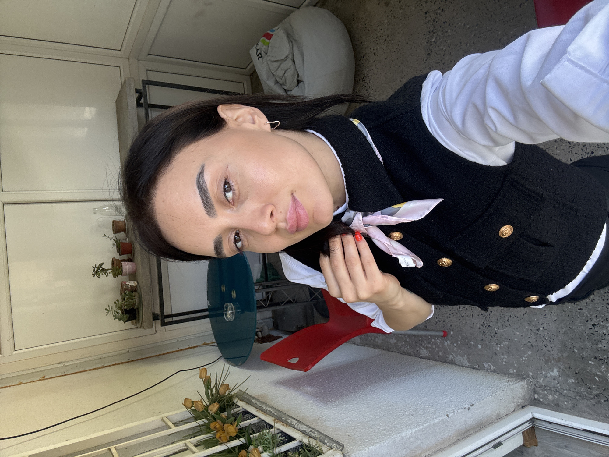 A young woman is depicted in a portrait-style photograph, rotated 90 degrees clockwise. She occupies the central and right portion of the frame, looking directly towards the camera with a soft expression and slightly parted pink lips. Her dark hair is pulled back, revealing a small silver heart-shaped earring on her visible ear. She has well-defined dark eyebrows and dark eyes.
She is dressed in a crisp white long-sleeved shirt layered under a black textured vest or jacket adorned with gold-colored buttons. A light-colored scarf with a subtle pattern, possibly silk, is tied around her neck. Her left hand, with red nail polish, is near her chest, gently touching the scarf.
The background suggests an outdoor or semi-enclosed patio or balcony setting. To the left, several small potted plants, including succulents, sit on a shelf against a light-colored wall or window structure. Further left, a white framework resembling window panes or dividers is visible. To the right and below her, the ground appears to be made of grey concrete or a similar rough surface. A white beanbag or rolled blanket is in the upper right corner. A red plastic chair is partially visible to her left. In the bottom left, a white low wall or platform is present, below which a metal grate holds a cluster of dried yellow flowers, possibly tulips, and some greenery. A dark electrical cable runs across the white surface. A teal or blue circular object is partially seen behind her.
The lighting appears to be natural daylight, indicating the photo was taken during the day. There is no visible text.