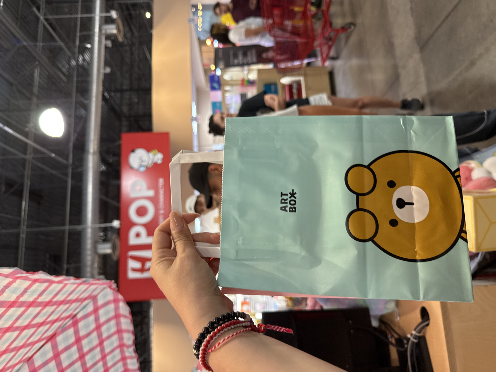 The image shows a person holding a light blue "ART BOX" shopping bag with a bear cartoon on it. The person is wearing multiple bracelets. The background includes a store setting with shelves and people, some with shopping carts. A red sign with "POP" and a cartoon character is also visible. The location is Orlando, United States, based on the ART BOX store presence in that city.