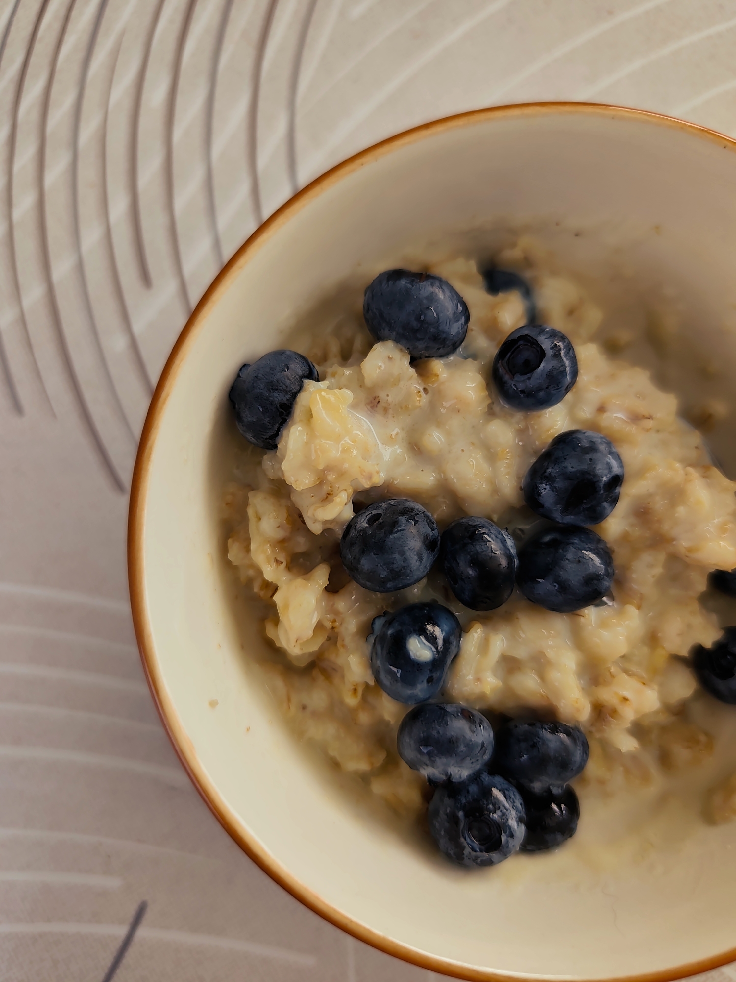 A close-up, overhead shot captures a bowl of oatmeal topped with fresh blueberries. The oatmeal appears creamy and slightly textured, with individual oat flakes visible. Several plump, dark blue blueberries are scattered across the surface of the oatmeal, some whole and others partially submerged. The bowl itself has a light-colored interior and a reddish-brown rim. It sits on a surface with a patterned texture, characterized by alternating light and dark wavy lines. The lighting is soft, creating subtle shadows and highlighting the glossy sheen of the blueberries. The scene is devoid of any people or activity, focusing solely on the food and its presentation. It is not possible to determine the specific location in the city of Durlești, Moldova, from the visual information provided in the image.