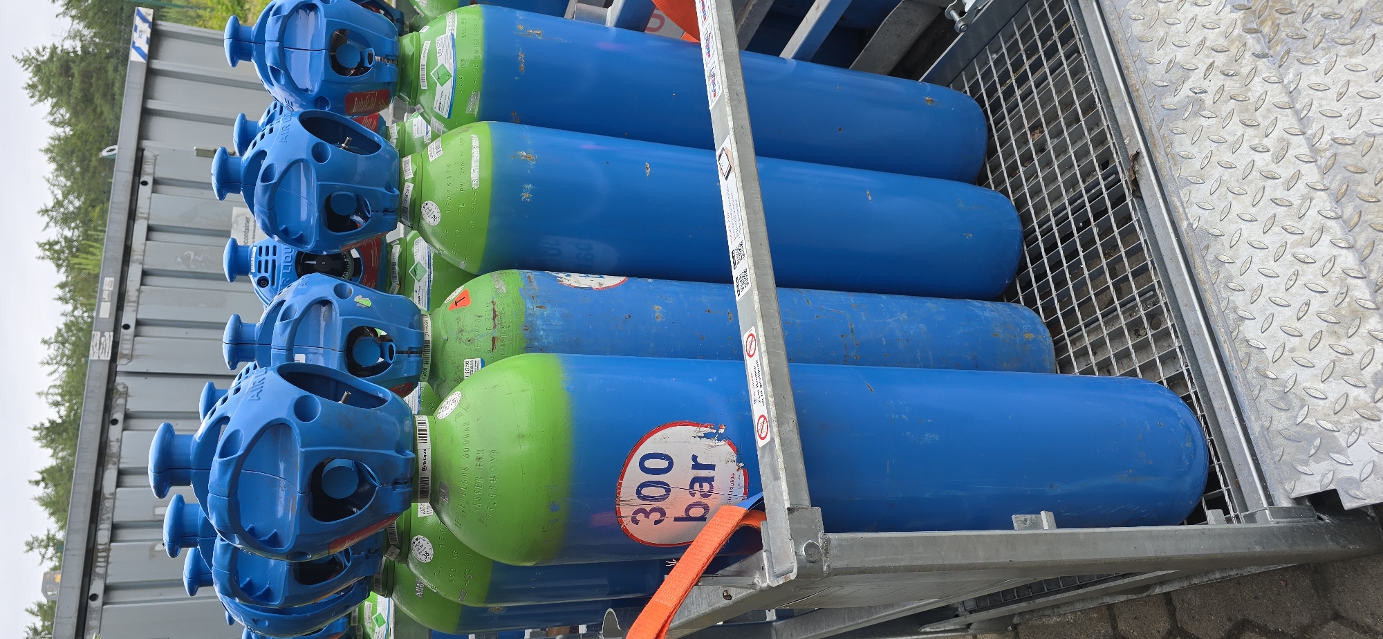 In Haren (Ems), Germany, a metallic cage-like structure holds a vertical arrangement of gas cylinders. The cylinders are predominantly blue with green tops. One prominent cylinder in the foreground displays a white circular label with "300 bar" printed in bold blue letters. The cylinders appear to be stored outdoors, with a grey industrial-style building and green trees visible in the background. An orange strap is attached to the cage structure, hinting at a loading or securing mechanism.