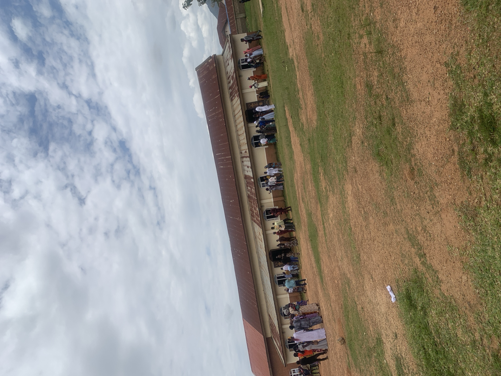 The image shows a group of people gathered outside a long, single-story building with a rusted, corrugated metal roof. The building has a light-colored facade and several arched windows. The ground in front of the building is a mix of dry, reddish-brown dirt and patchy green grass. In the foreground, a piece of white paper is crumpled on the ground. The sky is overcast with a mix of white and gray clouds. The people are dressed in various colorful clothing, and some appear to be standing in lines, possibly waiting to enter the building. The scene appears to be outdoors in a rural or semi-rural setting. The location is identified as Duk, Nigeria.
