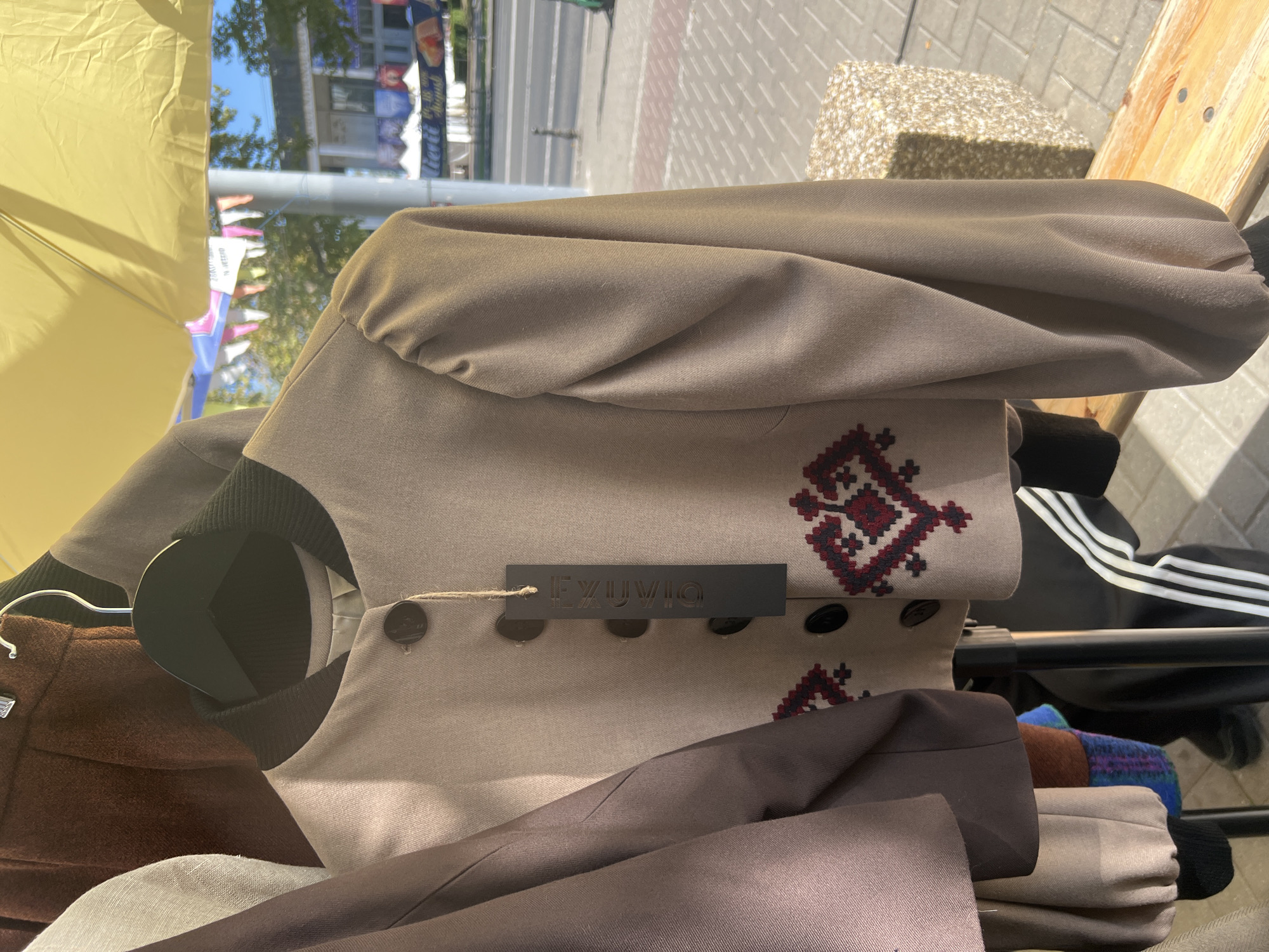 The image shows a close-up of a light brown, cropped bomber jacket with gathered sleeves, hanging on a black hanger. The jacket has a black ribbed collar and cuffs, and features four dark buttons down the front. A black tag with embossed white text, possibly a brand name "EXYUVIQ," is attached to the jacket by a string. A traditional embroidered pattern in red and dark blue is visible on the lower front of the jacket. To the left of the jacket, parts of other garments are visible, including a brown coat or skirt and a light gray garment. In the background, a yellow umbrella is visible, and beyond that, a street scene with buildings, trees, and pedestrians can be discerned. This scene is likely from a street market or outdoor event in Chișinău, Moldova, possibly near a building with flags or banners, suggesting a public area with commercial activity. The sun is shining, casting shadows on the paved ground and a wooden bench in the foreground.
