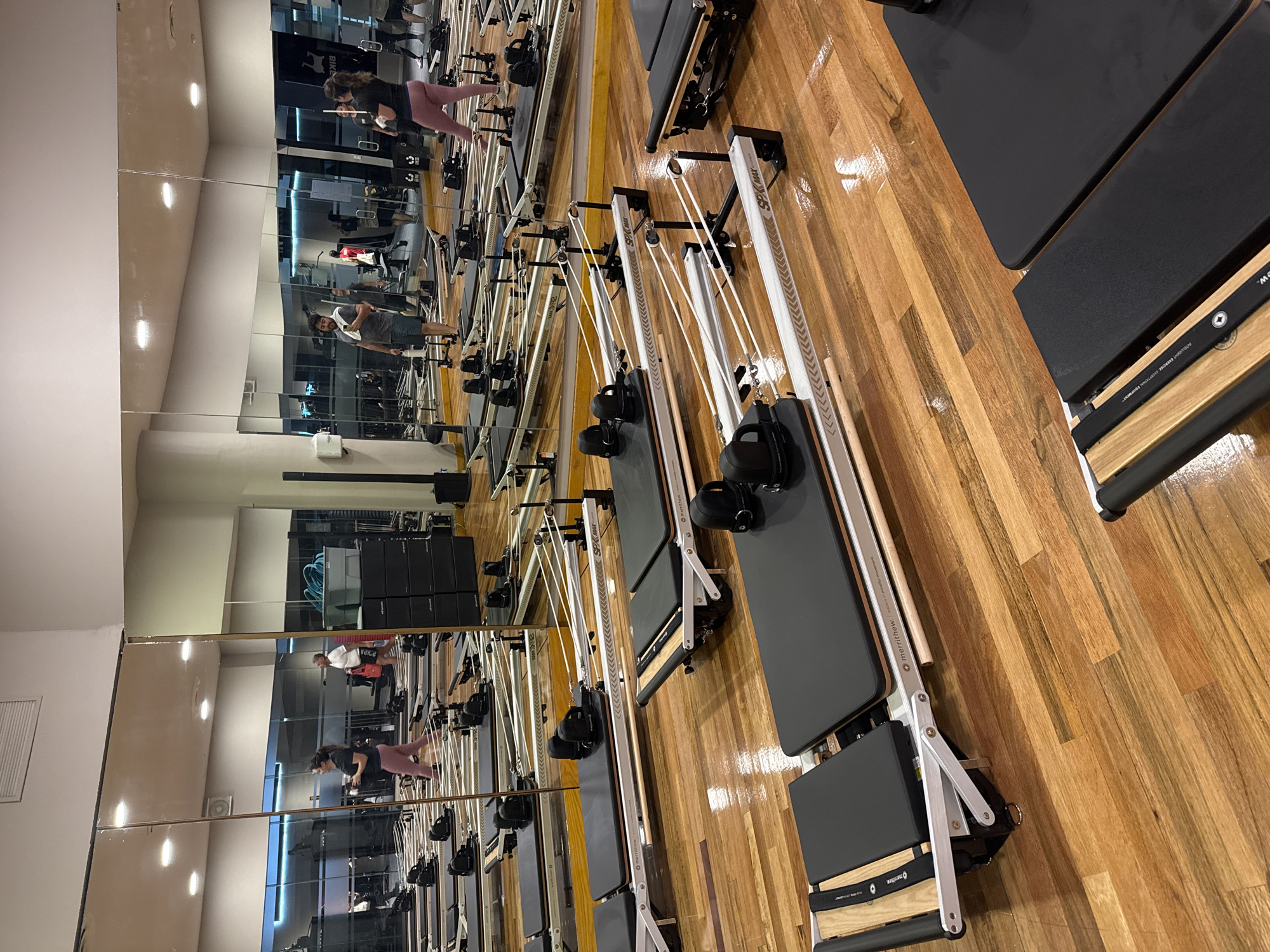 The image depicts the interior of a Pilates studio. Several Pilates reformer machines are positioned on a wooden floor, lined up in a row. These machines are silver with black padding and various adjustable straps and pulleys. Mirrored walls reflect the studio, showing images of people exercising on similar machines. One individual is wearing grey clothes with a white towel, while another is wearing pink pants and black clothes. The studio appears to be well-lit and organized. The text visible on the machines suggests the brand is "merrithew. intelligent exercise exceptional equipment." The location of the image is not confirmed, but given the clues it may be within Puebla, Mexico.
