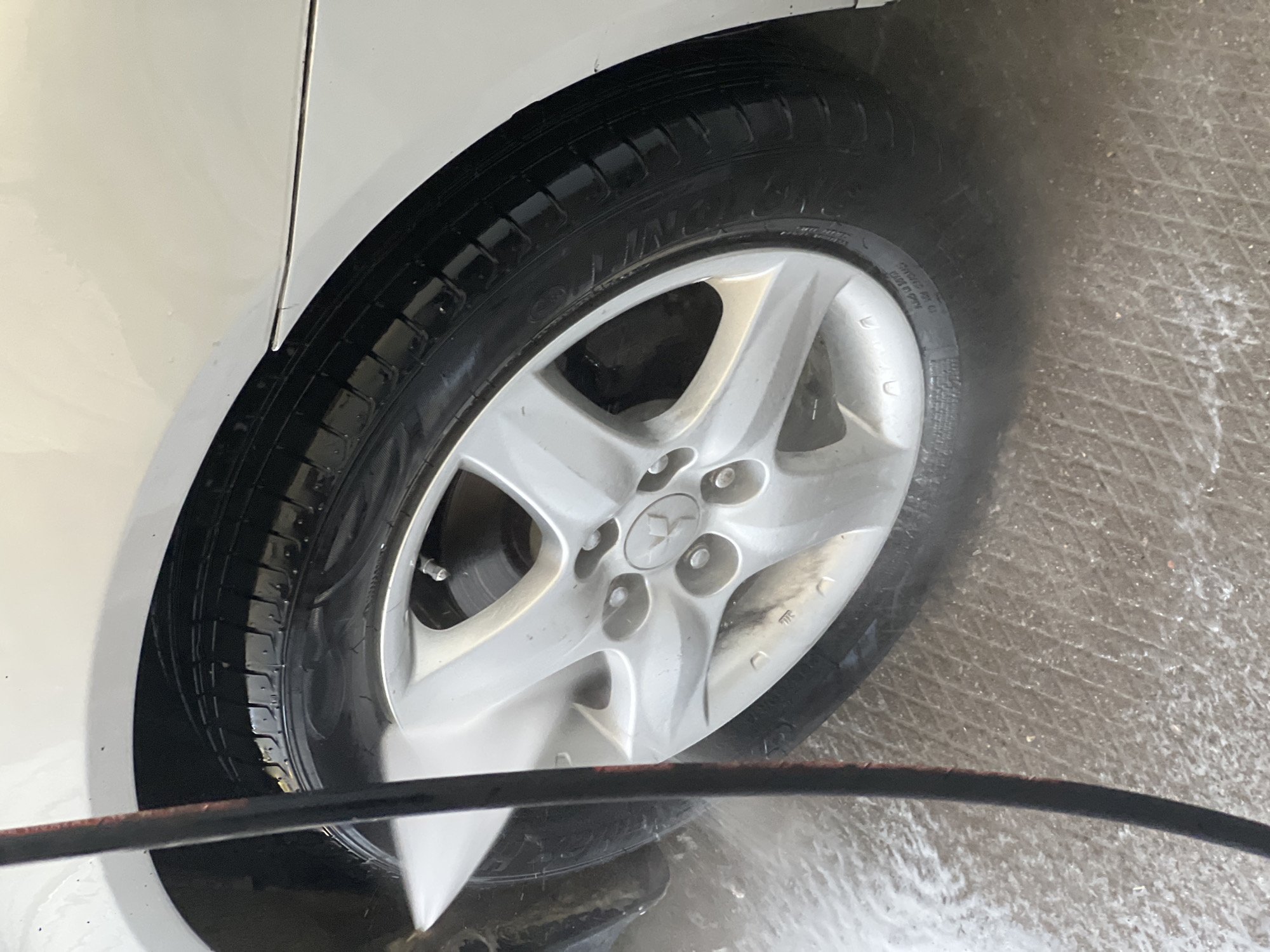 In the city of Shchelkino, Ukraine, a white car is being washed. The image focuses on the front passenger-side wheel and tire. A black hose, likely connected to a pressure washer, is visible in the foreground, spraying water onto the tire and rim. Water is actively splashing around the wheel, indicating the washing process is underway. The car's body, painted in a bright white, is partially visible above the wheel arch. The ground beneath the car appears to be a textured concrete surface, wet from the washing.