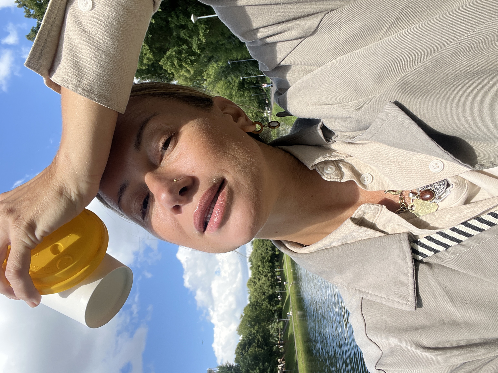 This is a close-up, outdoor, selfie-style photograph of a woman. She is holding a white paper cup with a yellow lid in her right hand. The woman has her left arm raised, resting her hand on her forehead, shielding her eyes from the sun. She is looking towards the camera with a gentle smile.
She is wearing a light-colored button-up shirt, possibly a trench coat, over a lighter linen top. She has subtle makeup on, with glossy lips and a nose stud. She is also wearing earrings and a charm bracelet with multiple pendants, including one that appears to say "YORK".
The background suggests a park setting with a body of water, likely a river or lake, to the right. Lush green trees line the opposite bank, and a clear blue sky with scattered white clouds is visible above. The sunlight indicates it is daytime. The overall mood of the image is relaxed and pleasant.
No visible text is discernible beyond the word "YORK" on a charm. The context provided indicates the location is Moscow, Russia.