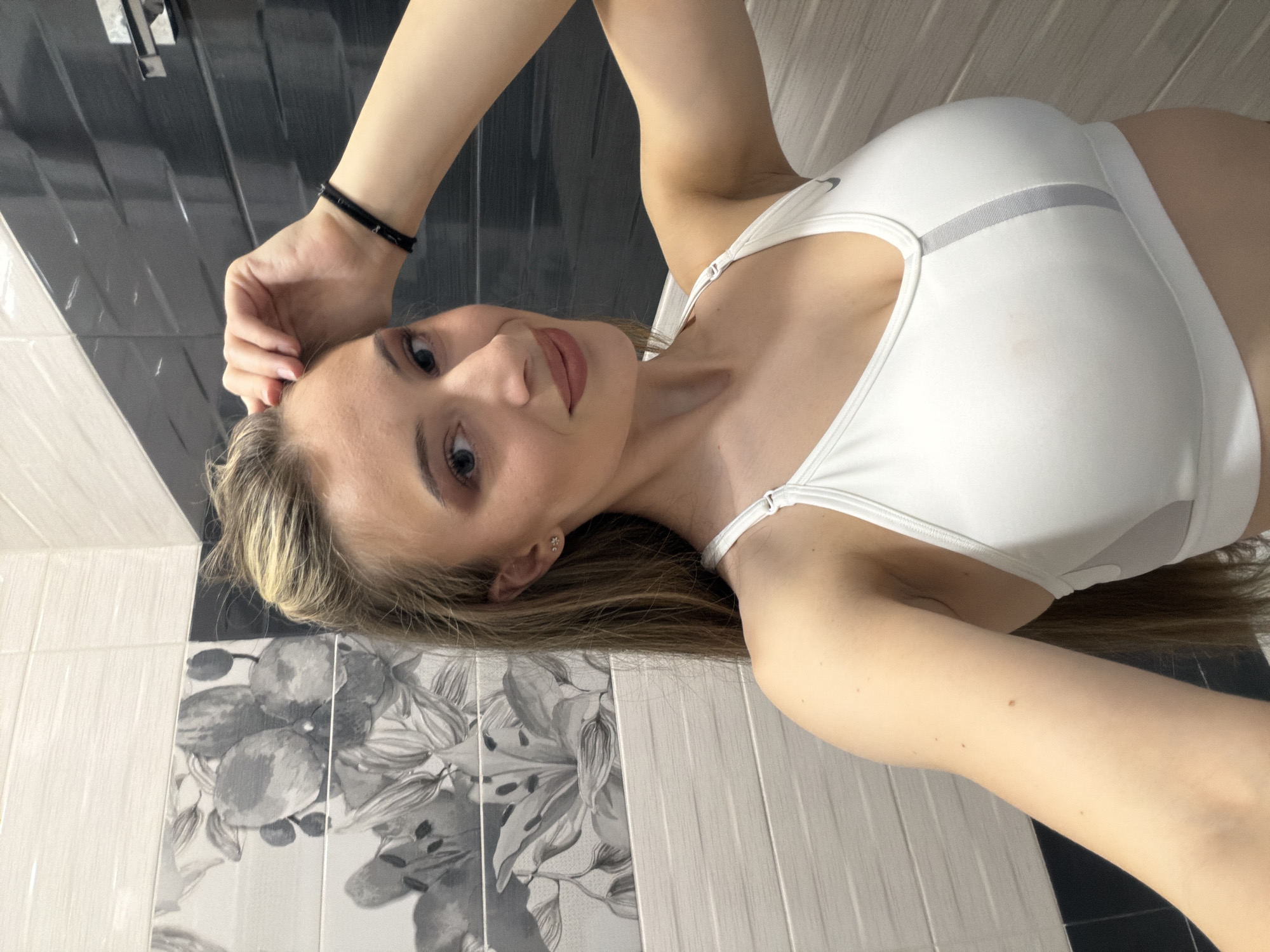 A woman is posing for a selfie in a bathroom. She is wearing a white sports bra and has her left hand resting on her head. The background consists of patterned tiles and plain black and white tiles. No information indicates the location is in Plovdiv, Bulgaria.