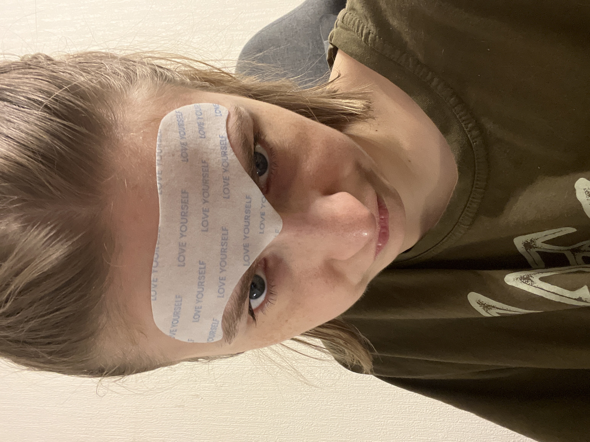 A portrait captures a person with fair skin, light brown hair, and blue eyes, oriented at an oblique angle. A rectangular patch, printed with the words "LOVE YOURSELF," is adhered to their forehead, partially covering one eyebrow. The person is wearing a dark green T-shirt with a faded white graphic on the front. The background consists of a white wall and a gray area, possibly a pillow or headrest. The location is not possible to determine.