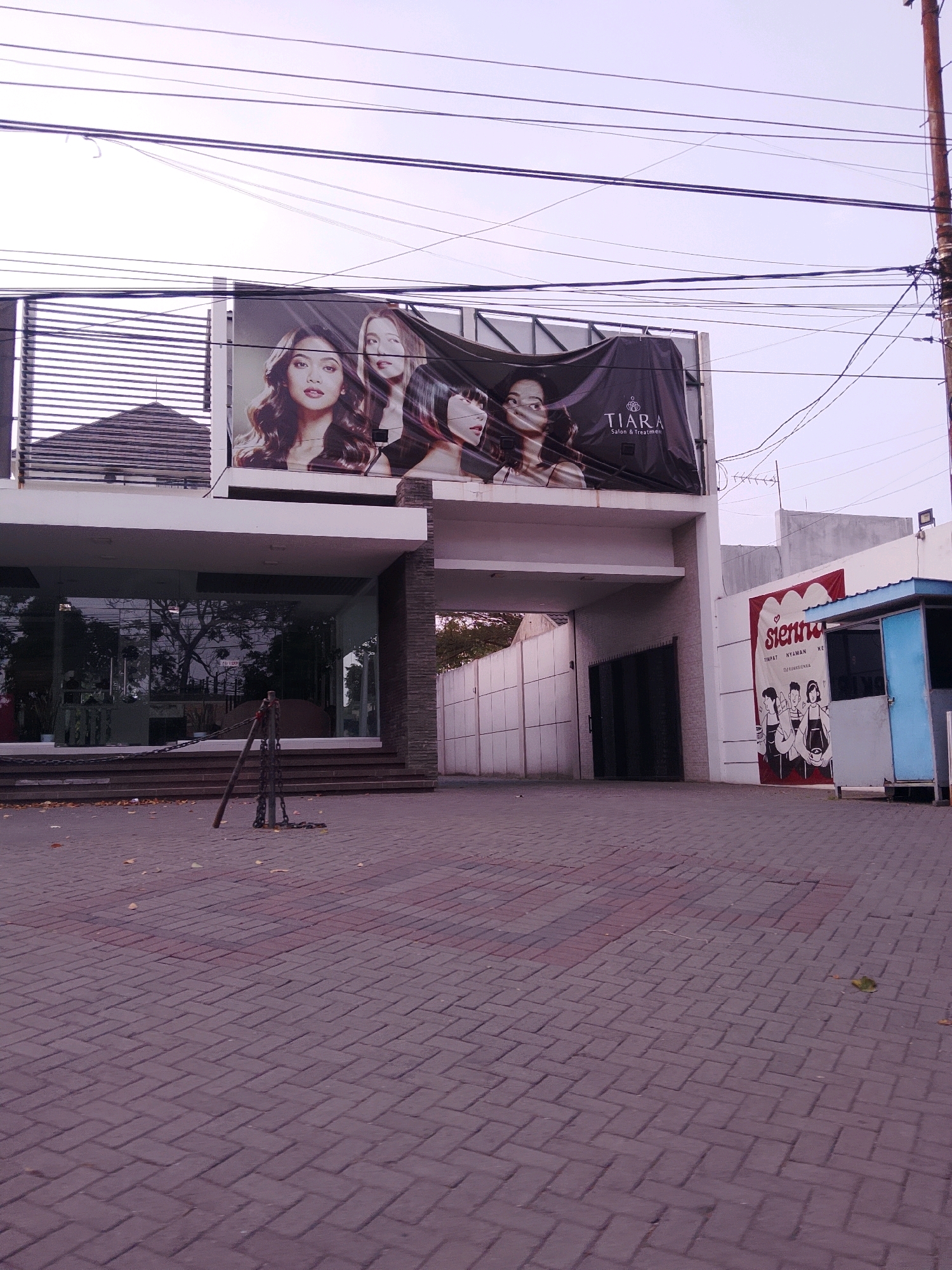 The image depicts the entrance to a building, likely a salon, in Malang, Indonesia. A large banner featuring three women with styled hair and the text "TIARA Salon & Treatment" is prominently displayed above the entrance. To the right of the building, another banner with cartoon-style drawings of people and the word "sienna" is visible. The ground in front of the building is paved with reddish-brown bricks laid in a herringbone pattern. Several electrical wires are strung across the sky, and a utility pole is visible on the right side of the image. A chain barrier is positioned in front of the building's entrance. The overall scene suggests a commercial establishment in an urban setting.