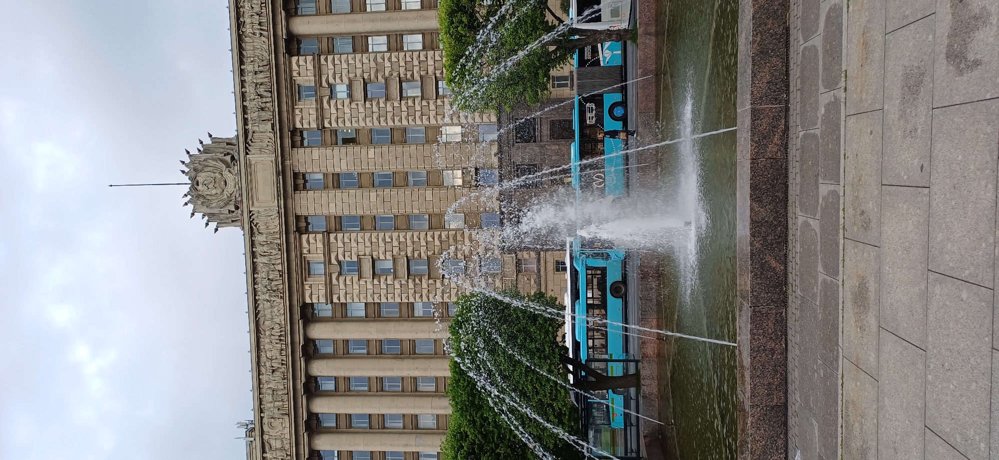 The image depicts a street scene in Saint Petersburg, Russia, with a prominent fountain in the foreground. The fountain features multiple jets of water spraying upwards into the air, creating a misty effect. Behind the fountain, a large, classical building with columns and intricate relief sculptures adorns its facade. The building has a grey stone finish and many windows. Two bright blue articulated buses are visible parked on the street in front of the building, with one person standing near the side of the bus. Lush green trees line the area between the fountain and the buses. The sky above is overcast with clouds. The foreground shows a stone-tiled pathway.