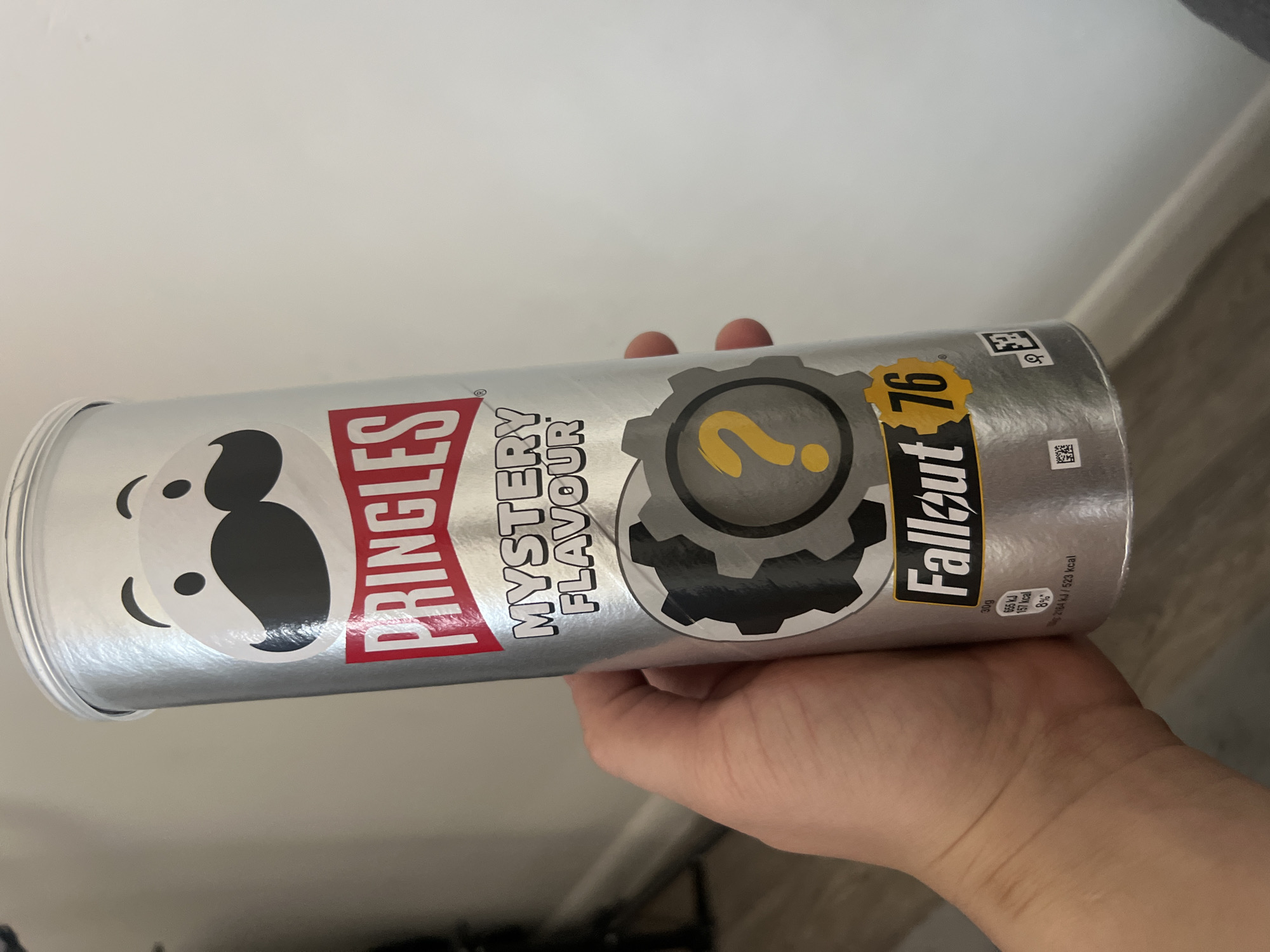 A hand holds a silver Pringles can. The can is labeled "Pringles Mystery Flavour" with a "Fallout 76" logo that includes a yellow question mark inside a gear. Nutritional information is printed near the bottom of the can. The background is a plain white wall. The image does not contain any information about Gorleston-on-Sea, United Kingdom.