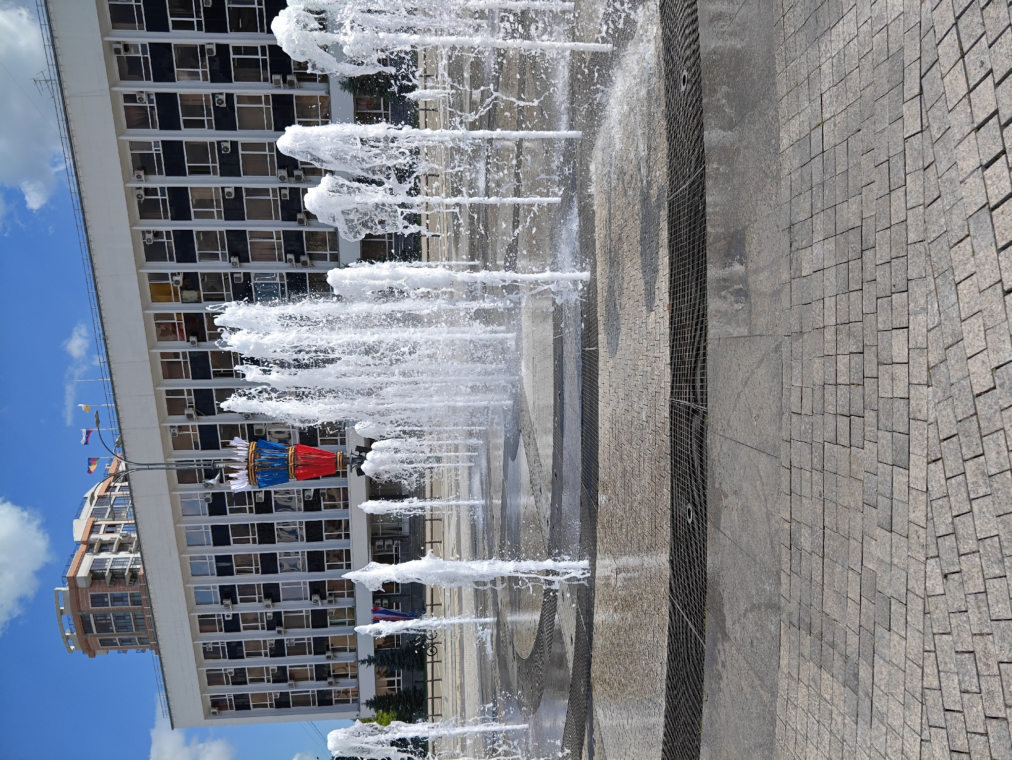 This is a photograph taken at the Fountain Square in Krasnodar, Russia, on a sunny day with a blue sky and scattered clouds. The foreground is paved with light-colored cobblestones arranged in a geometric pattern. In the mid-ground, a circular fountain is active, with numerous jets of water spraying upwards and outwards, creating a dynamic and lively scene. The fountain is surrounded by a paved area with metal grates for drainage. Behind the fountain, a large, multi-story building with numerous windows and air conditioning units is visible. The building has a white facade and a dark band running across its width. A taller, modern building with a curved top is situated to the left of the main building. Several flags are visible on poles in front of the building. Trees are planted along the edge of the paved area in front of the building. A bicycle is parked near the entrance of the building. The overall impression is of a public space designed for recreation and gathering, with a blend of modern and classic architecture.