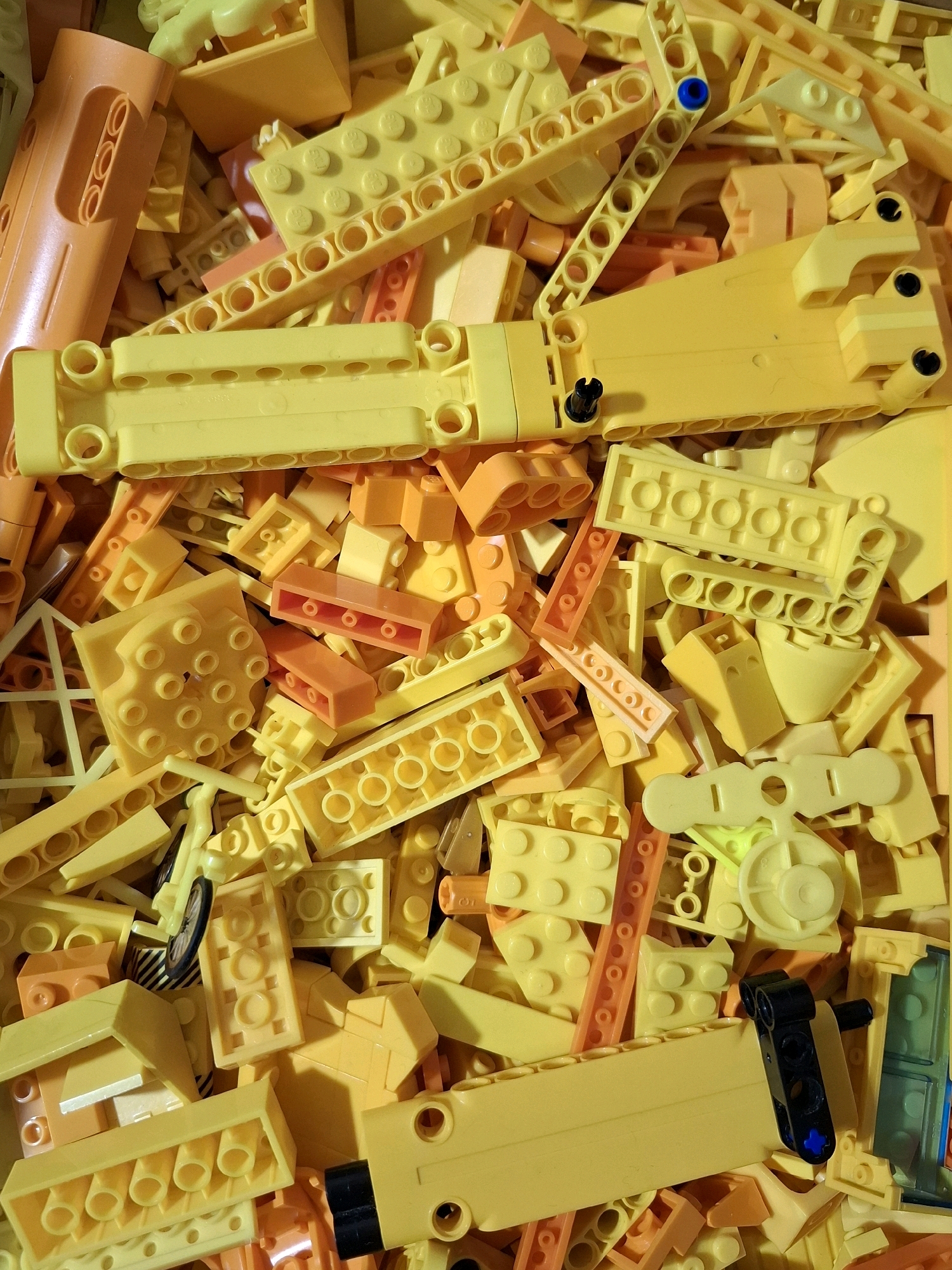A flat lay, top-down view of a large pile of yellow and orange plastic building blocks, resembling LEGOs. The blocks are of various shapes and sizes, including long rectangular bricks with multiple holes, smaller rectangular blocks, and some specialized pieces. There are also a few small black plastic connectors visible. The lighting casts shadows, creating a textured appearance. The image is an indoor close-up shot of a toy collection. There are no people, animals, or any elements that could indicate a specific location in the city of Tyumen, Russia.