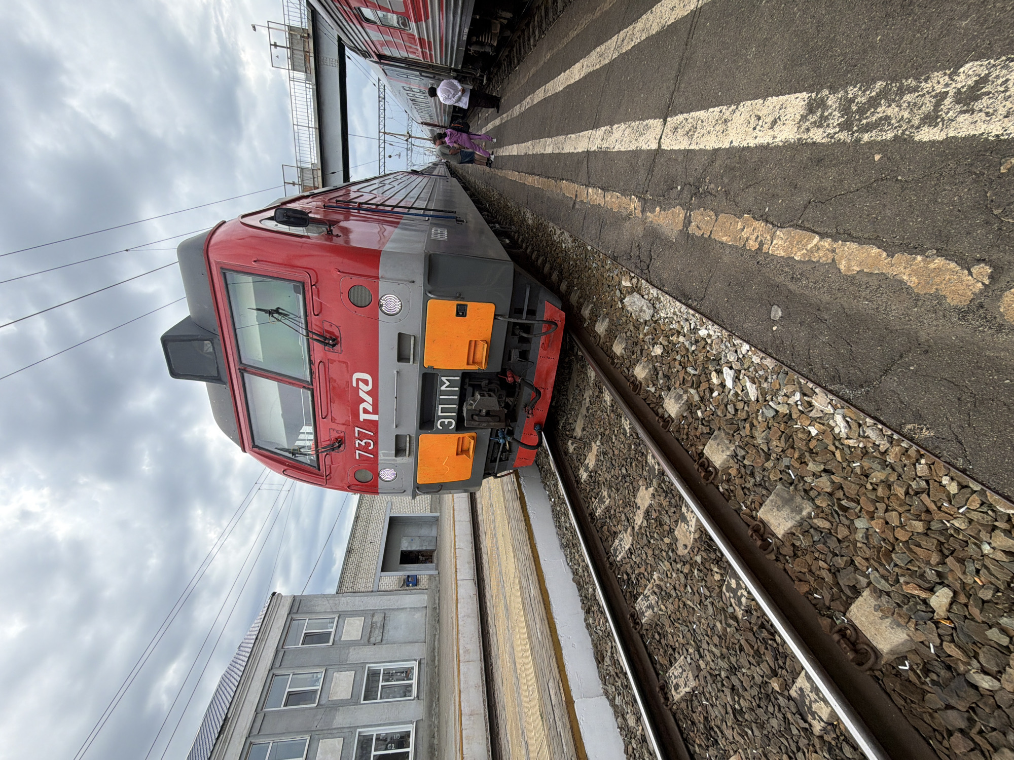 The image shows a red and gray train at a station, likely in Rtishchevo, Russia, given the context of the prompt. The train's front is in focus, displaying the number "737" and the Russian Railways logo. The train is positioned on tracks next to a concrete platform. In the background, a gray building with windows and a smaller brick structure are visible. Several people are standing on the platform near another train car. The sky is overcast with clouds. The ground on the platform is asphalt with painted yellow and white lines. The train tracks are surrounded by ballast.
